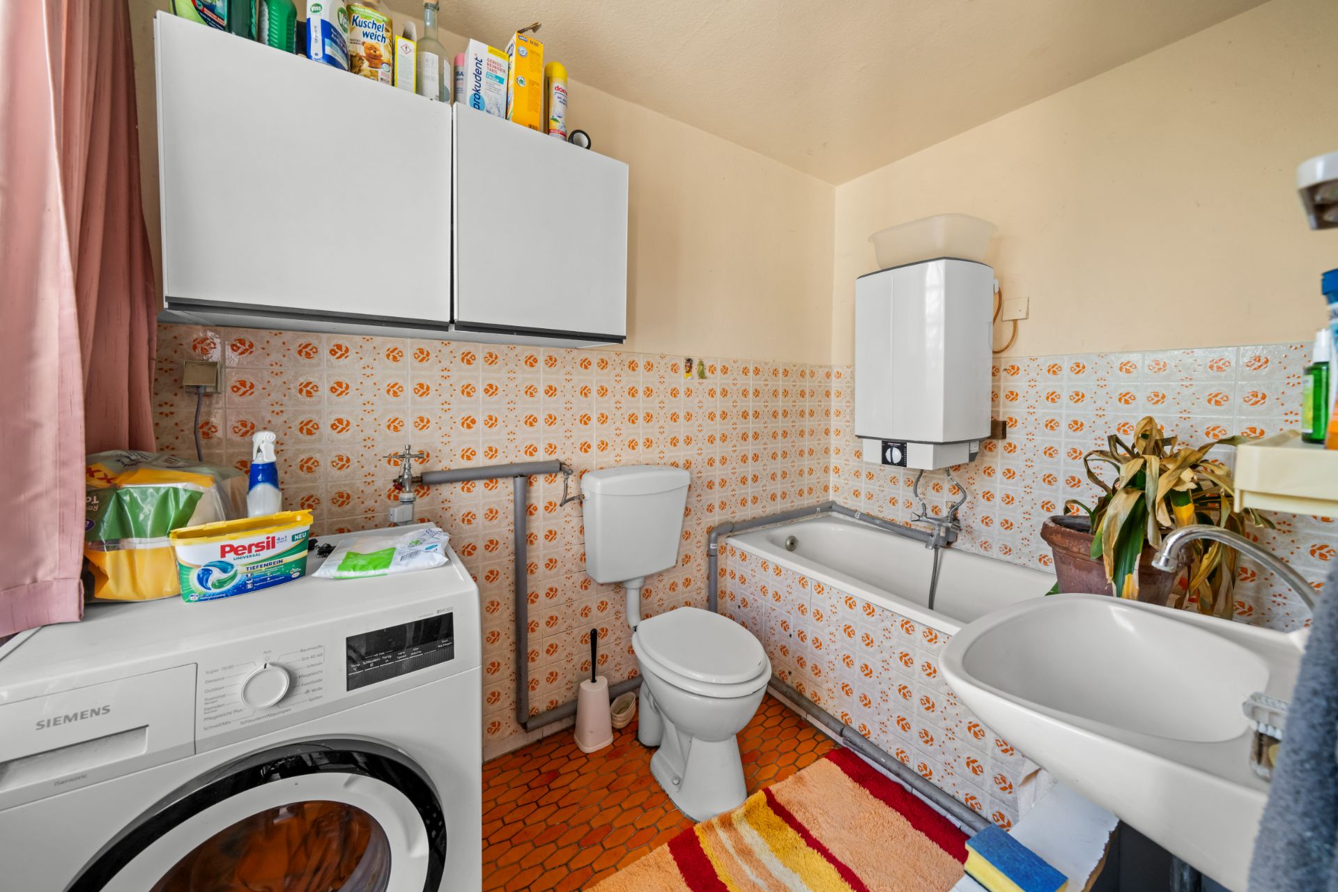 Badezimmer mit Waschmaschine, Toilette, Badewanne, Waschbecken und buntem Teppich.