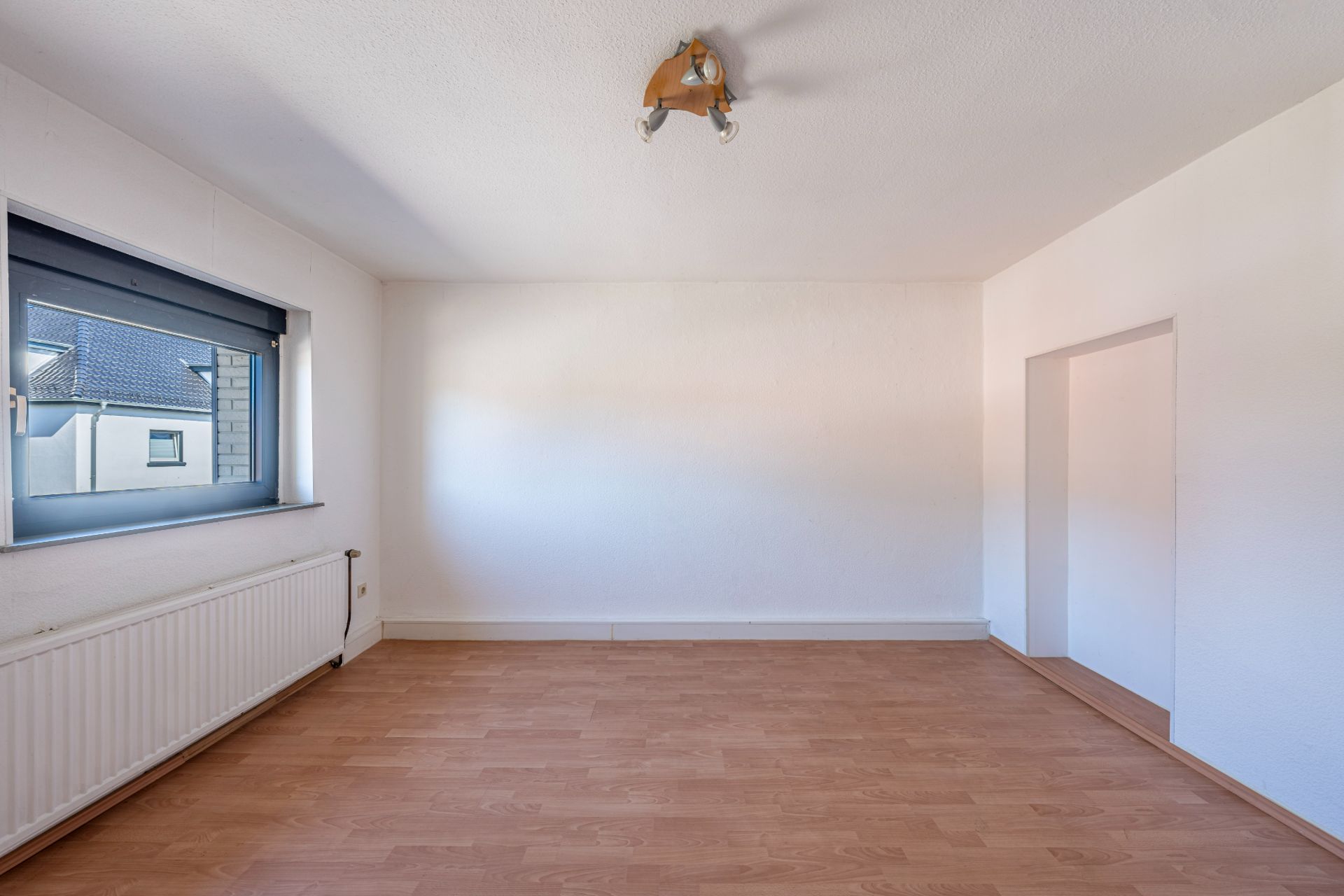 Helles, leeres Zimmer mit Holzfußboden, großem Fenster und Heizung an der Wand.