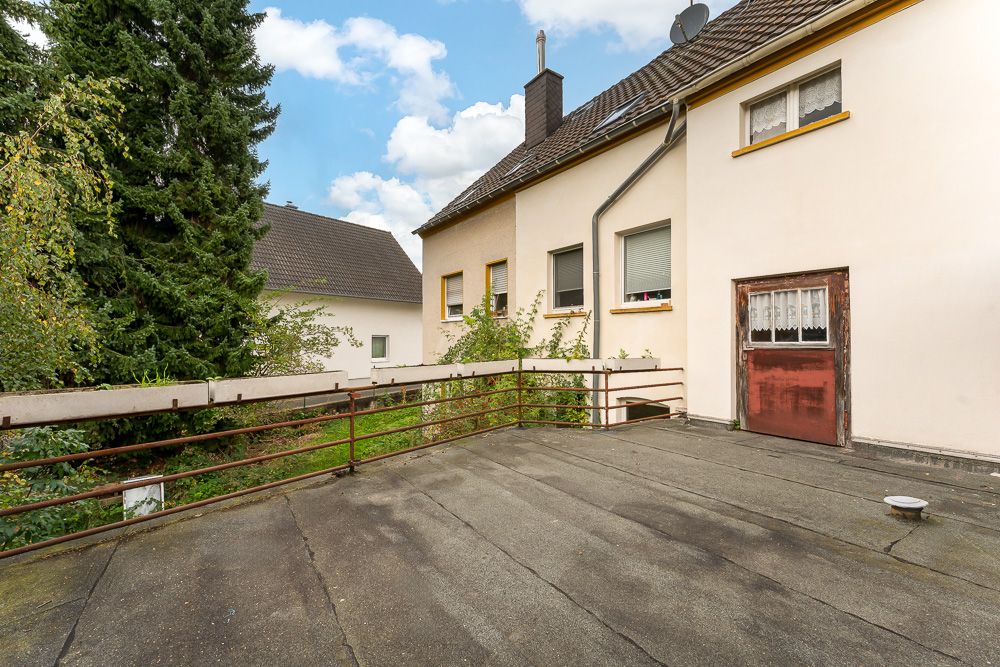 Große Terrasse mit Geländer an einem Wohnhaus, umgeben von Bäumen und Nachbarhäusern.