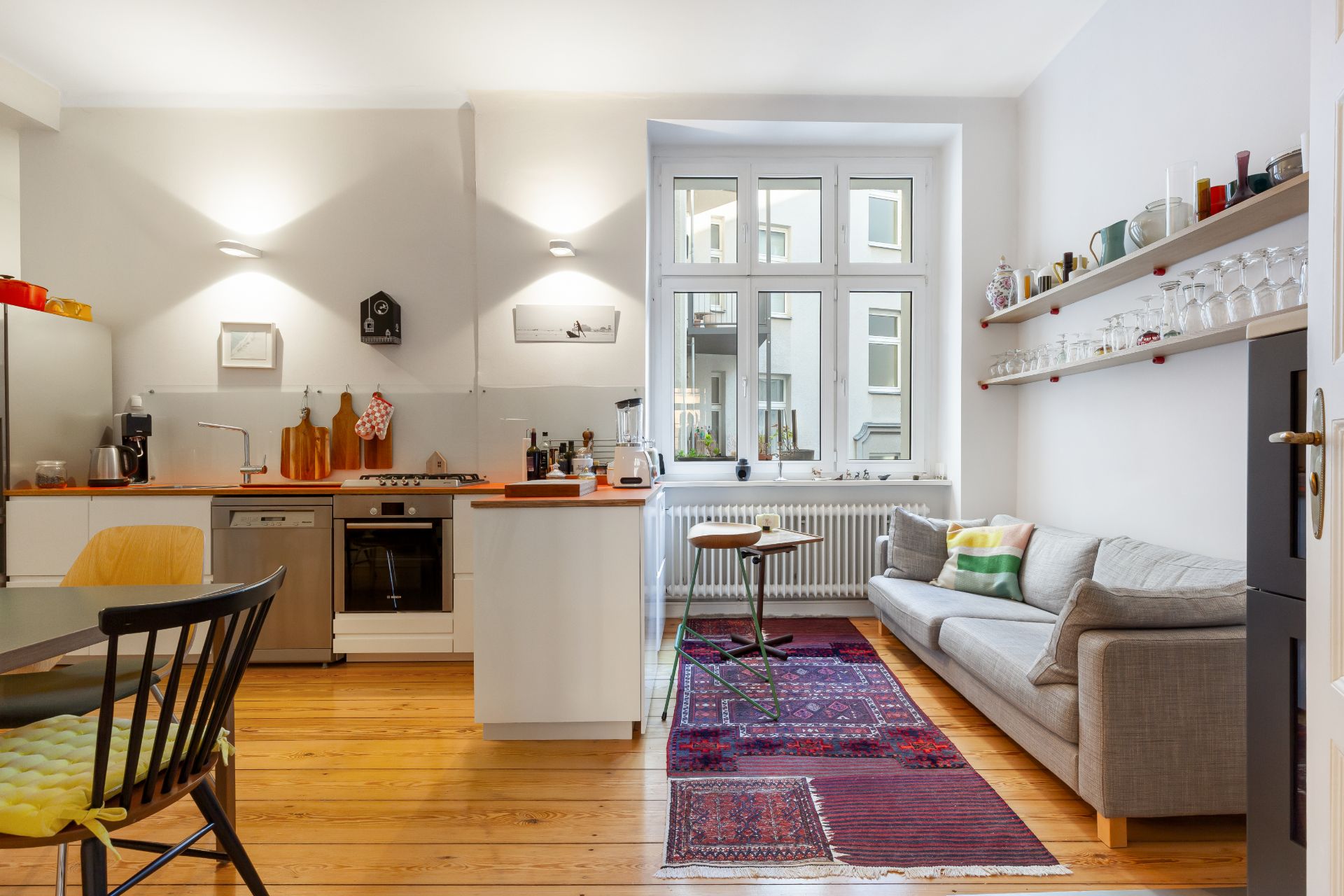 Offene Küche mit Esstisch, Sofa, Teppich und Fenster, Regal mit Gläsern an der Wand.