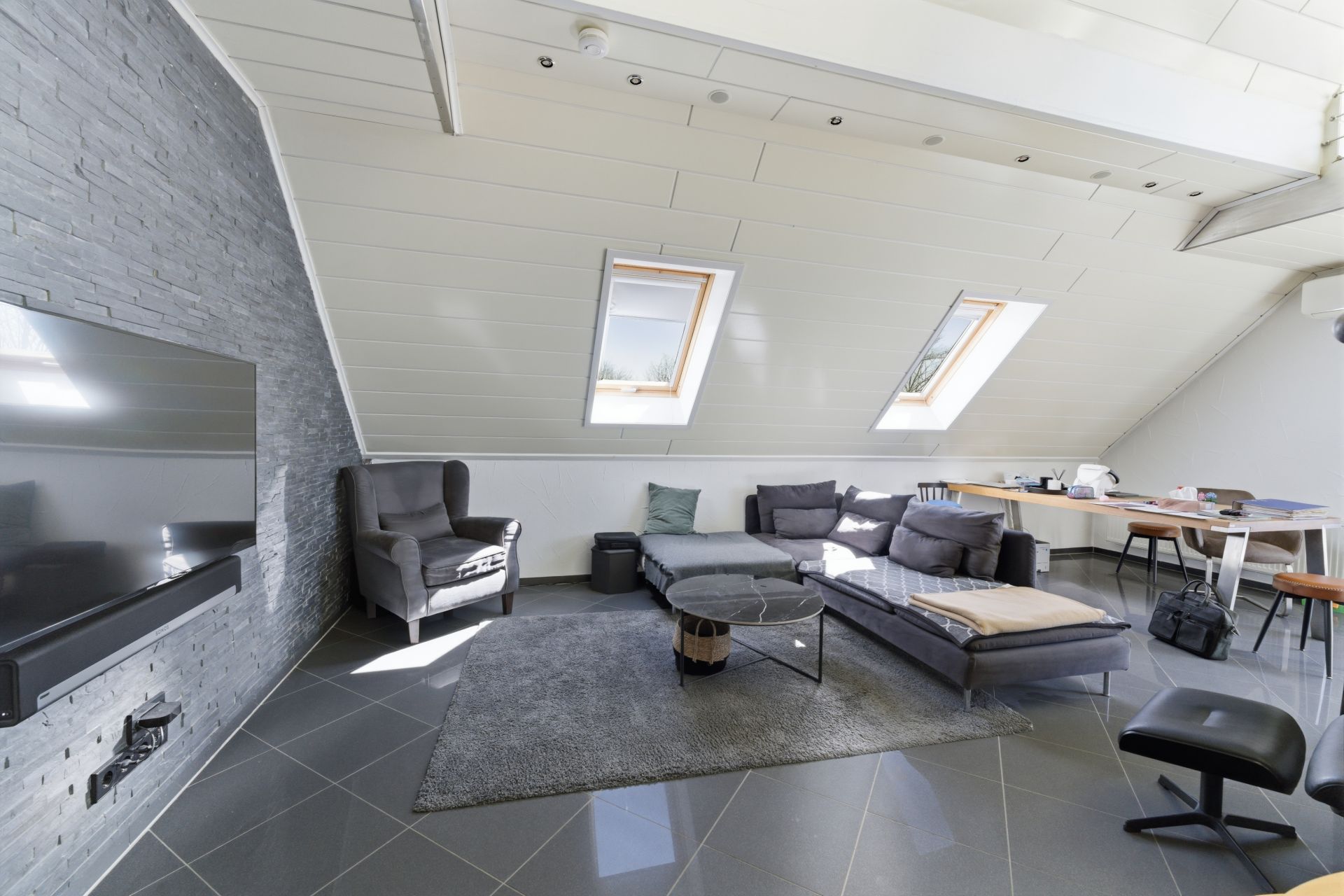Modernes Wohnzimmer mit Dachschräge, Sofa, Sesseln, Fernseher und zwei Dachfenstern.