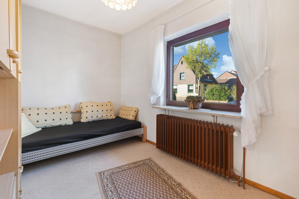 Helles Zimmer mit Sofa, Teppich, großem Fenster, Vorhängen und braunem Heizkörper.