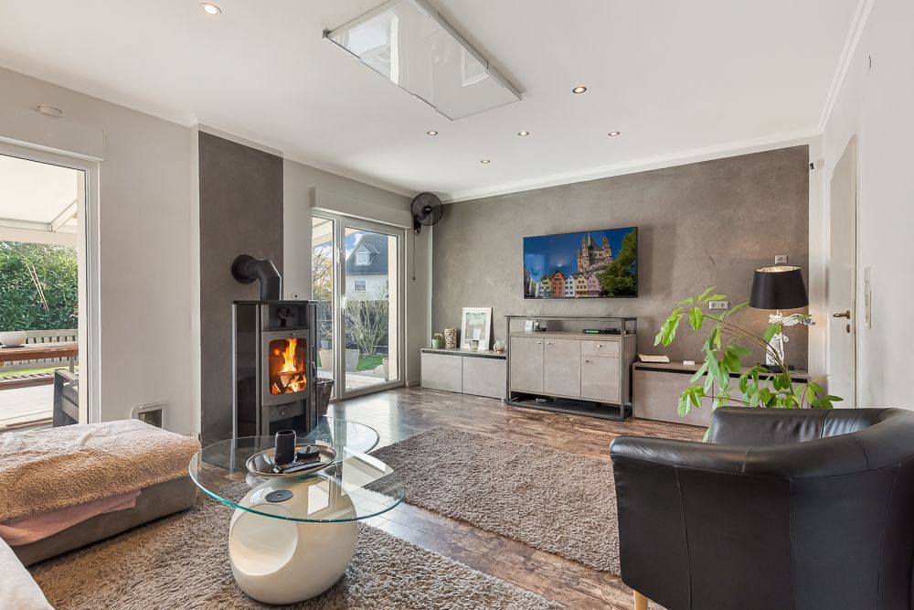 Modernes Wohnzimmer mit Kamin, Glastisch, TV und großen Fenstern zum Garten.