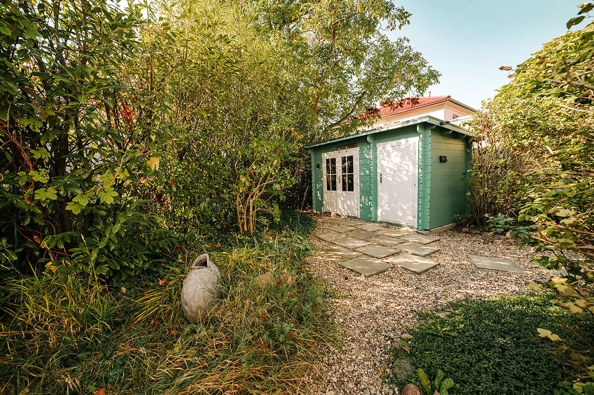Ein grünes Gartenhaus mit weißen Türen steht inmitten von Bäumen und Sträuchern.