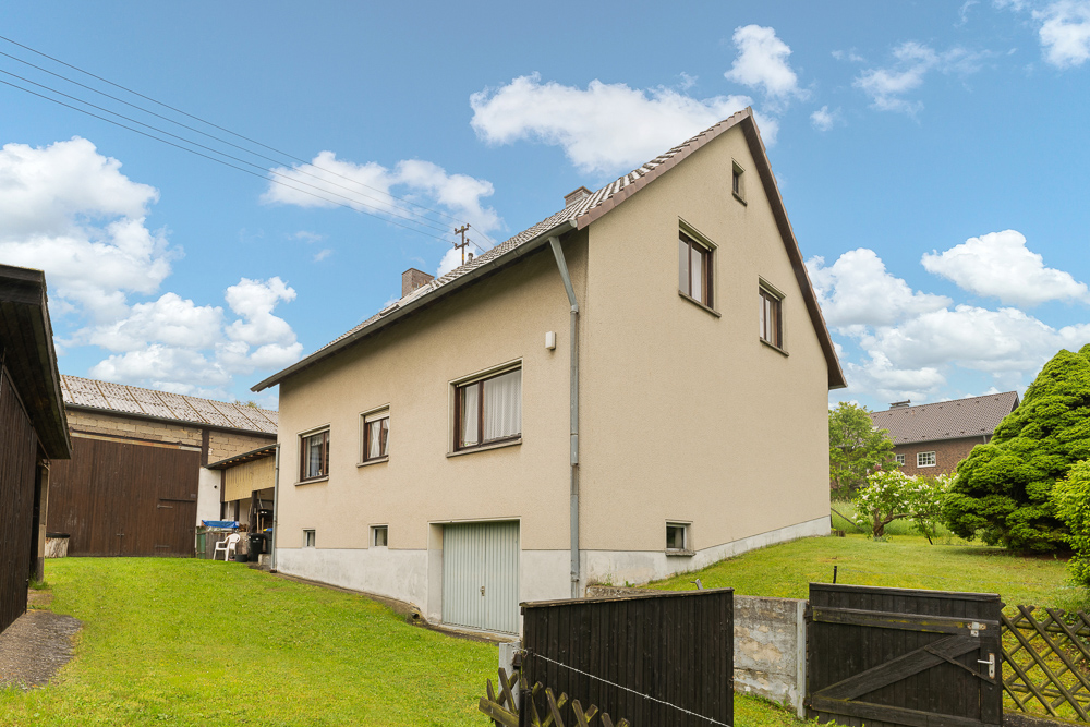 Beiges Wohnhaus mit Satteldach, mehreren Fenstern und Garage, umgeben von grünem Garten.