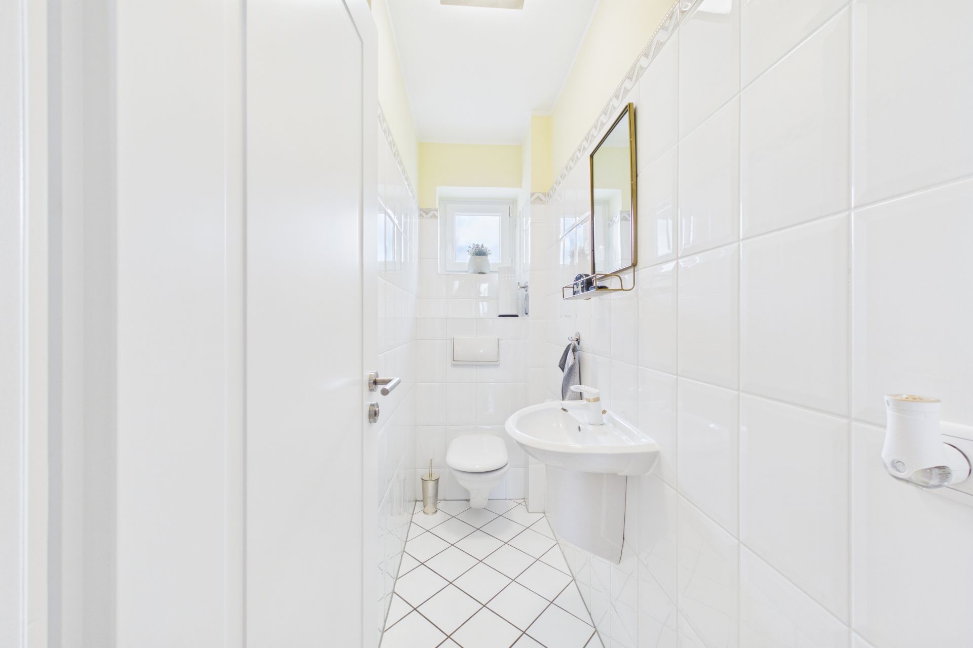 Helles, schmales Badezimmer mit Toilette, Waschbecken, Spiegel und Fenster am Ende.
