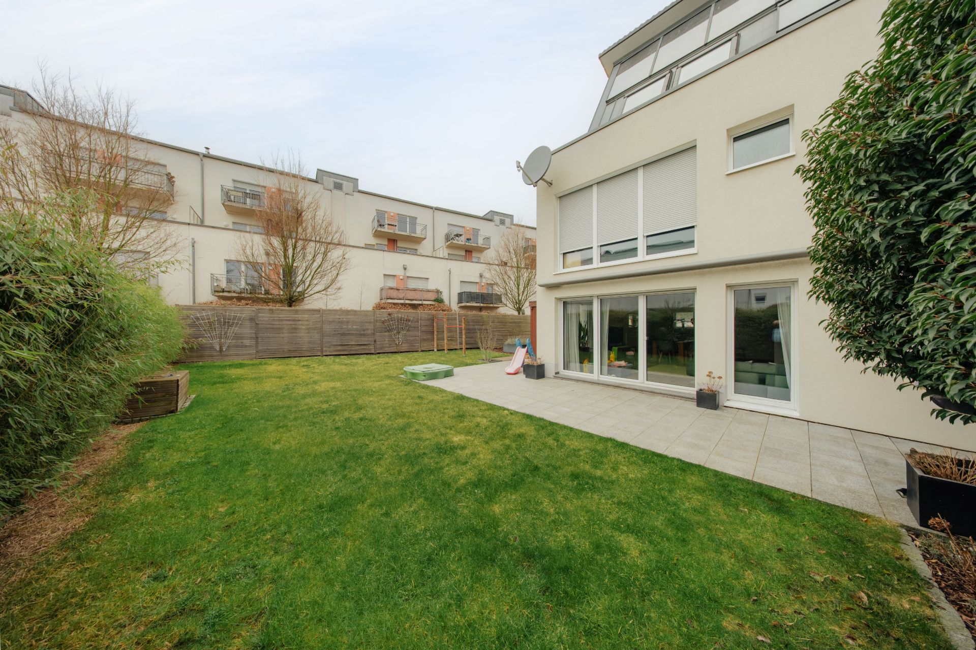 Grüner Garten mit Terrasse an einem modernen Wohnhaus, im Hintergrund weitere Häuser.