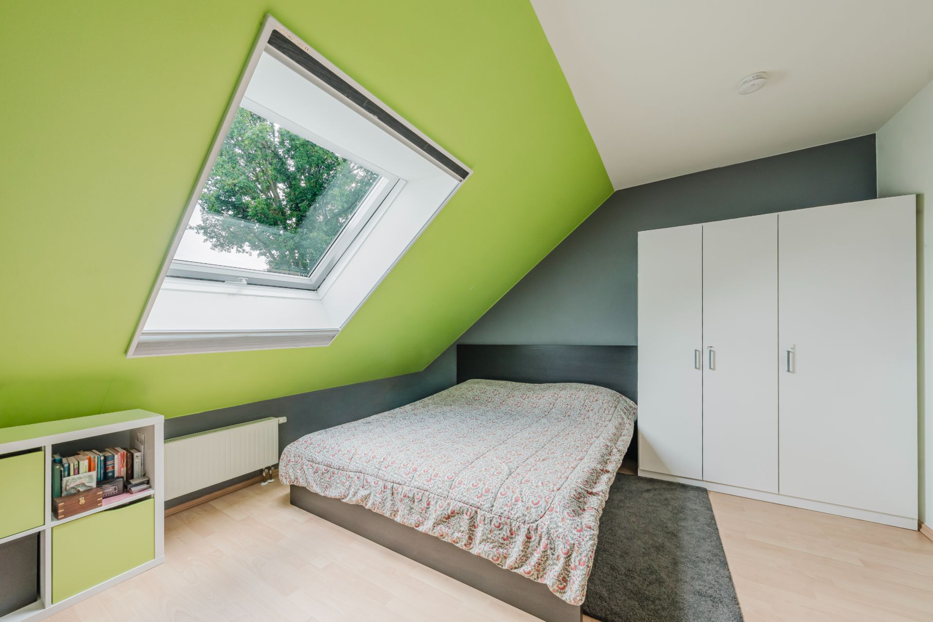Helles Schlafzimmer mit Dachfenster, Bett, weißem Kleiderschrank und Regal an einer grünen Wand.