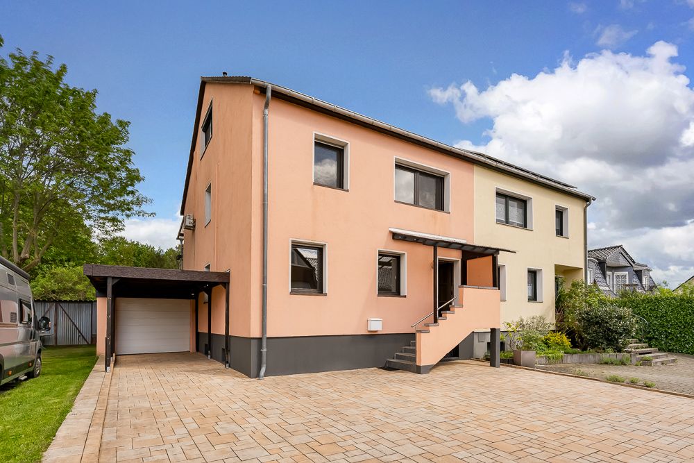 Zweistöckiges Wohnhaus mit Carport und gepflastertem Vorplatz, Eingangstreppe vorne.