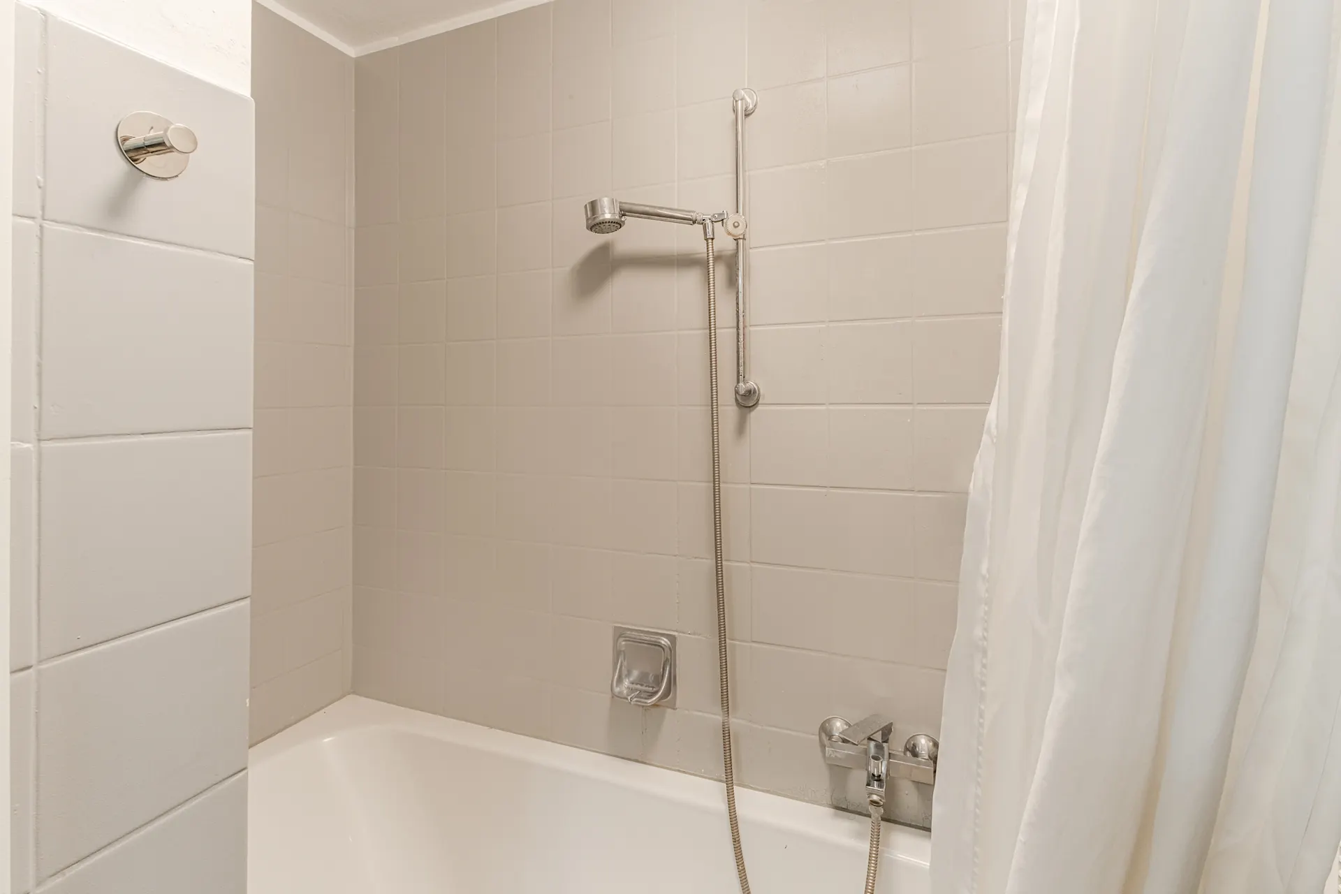Weiße Badewanne mit Duschvorhang und silberner Duscharmatur vor beigen Fliesen.