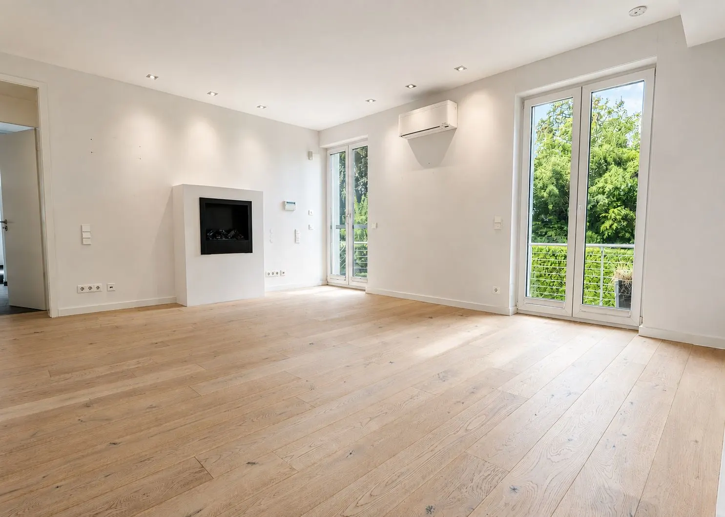 Helles, leeres Wohnzimmer mit Holzfußboden, Kamin und großen Fenstern mit Blick ins Grüne.