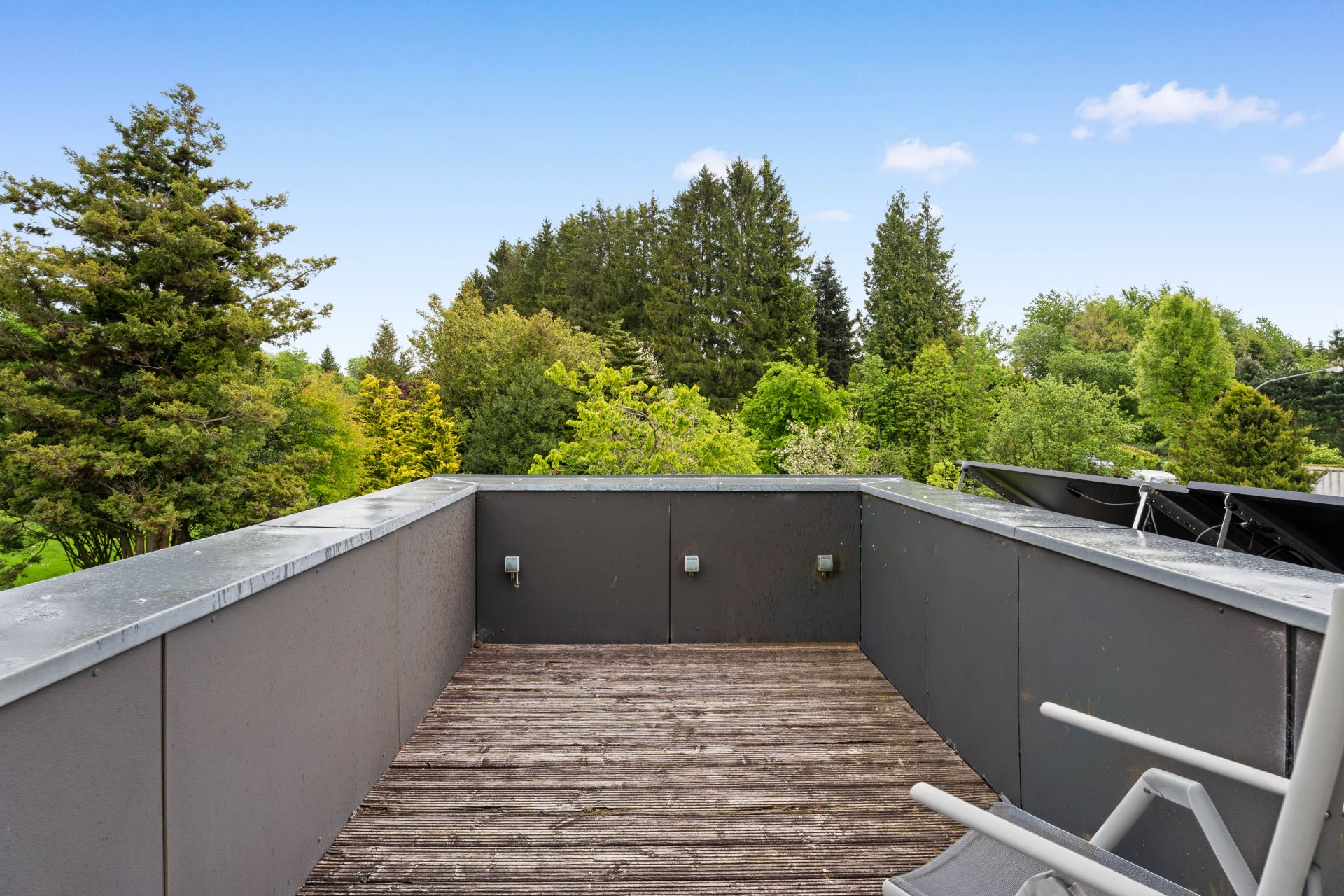 Kleine Dachterrasse mit Holzboden, grauen Wänden und Blick auf viele grüne Bäume.