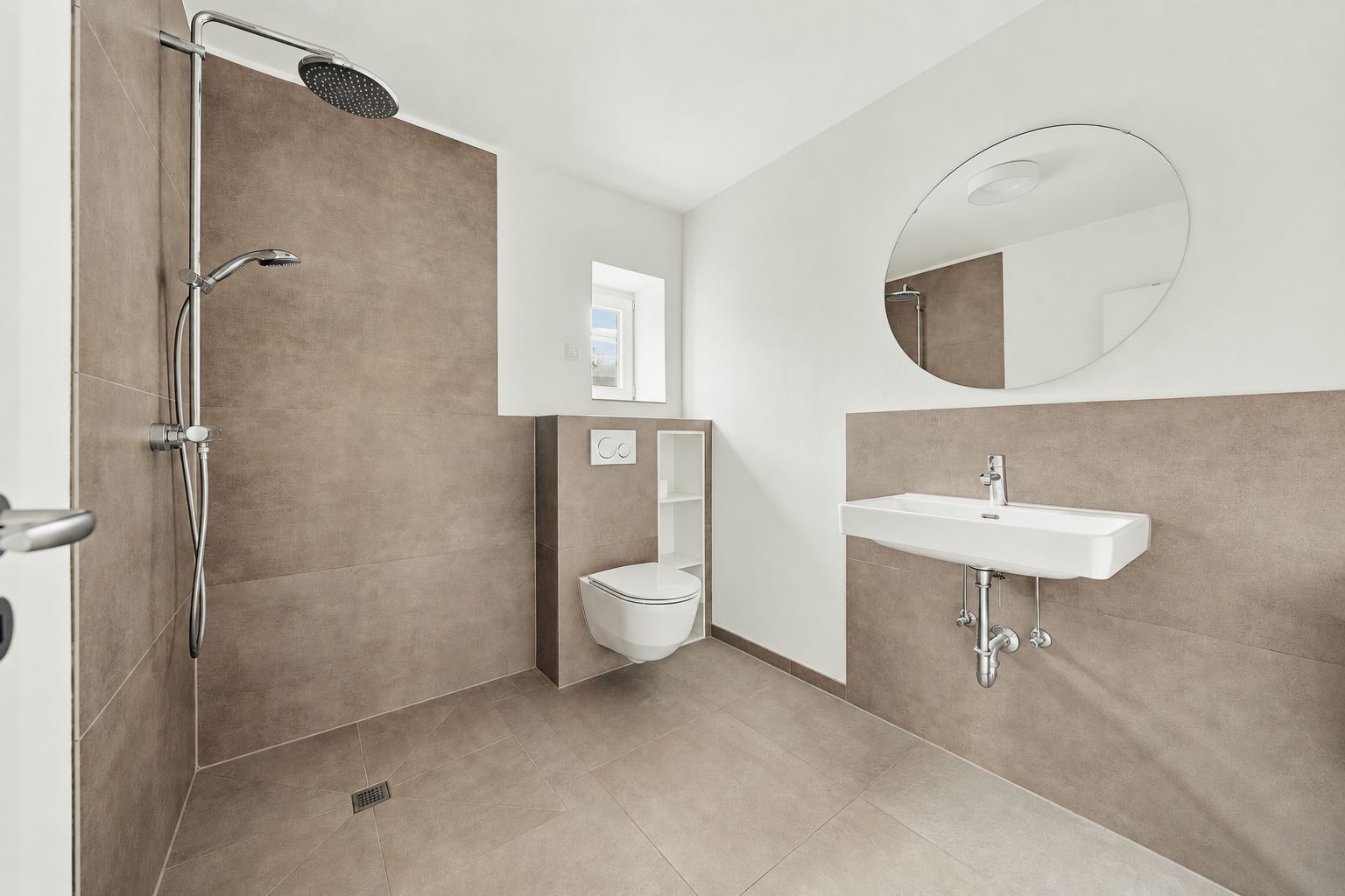 Modernes Badezimmer mit bodengleicher Dusche, Wandtoilette, Waschbecken und rundem Spiegel.