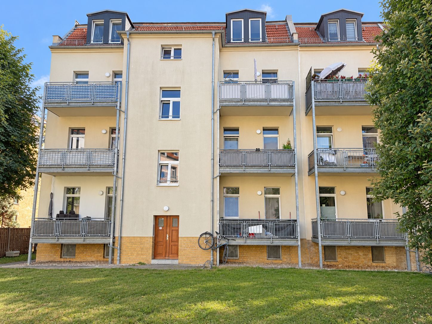 Mehrfamilienhaus mit Balkonen, gelber Fassade und grünem Vorgarten.