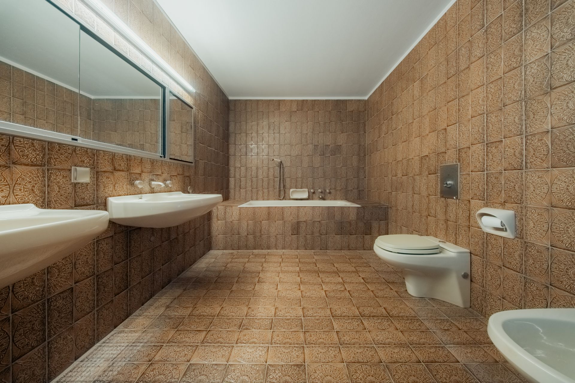 Badezimmer mit braunen Fliesen, zwei Waschbecken, Badewanne, Toilette und Bidet.
