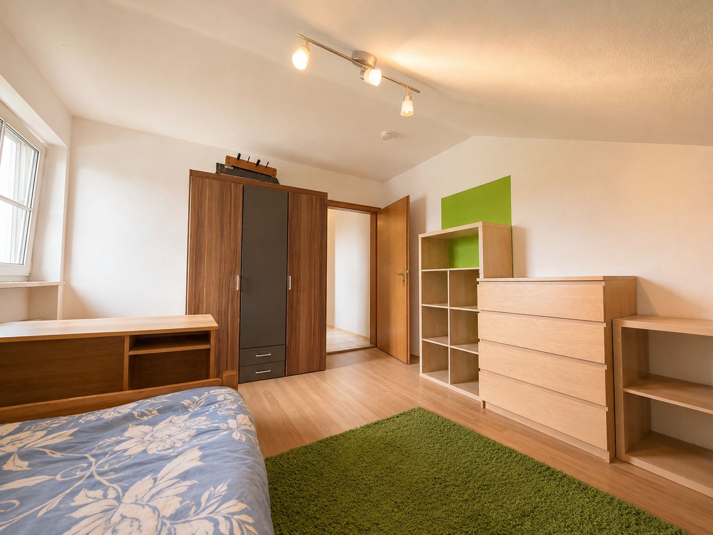 Helles Schlafzimmer mit Bett, Schrank, Kommode, Regal und grünem Teppich.