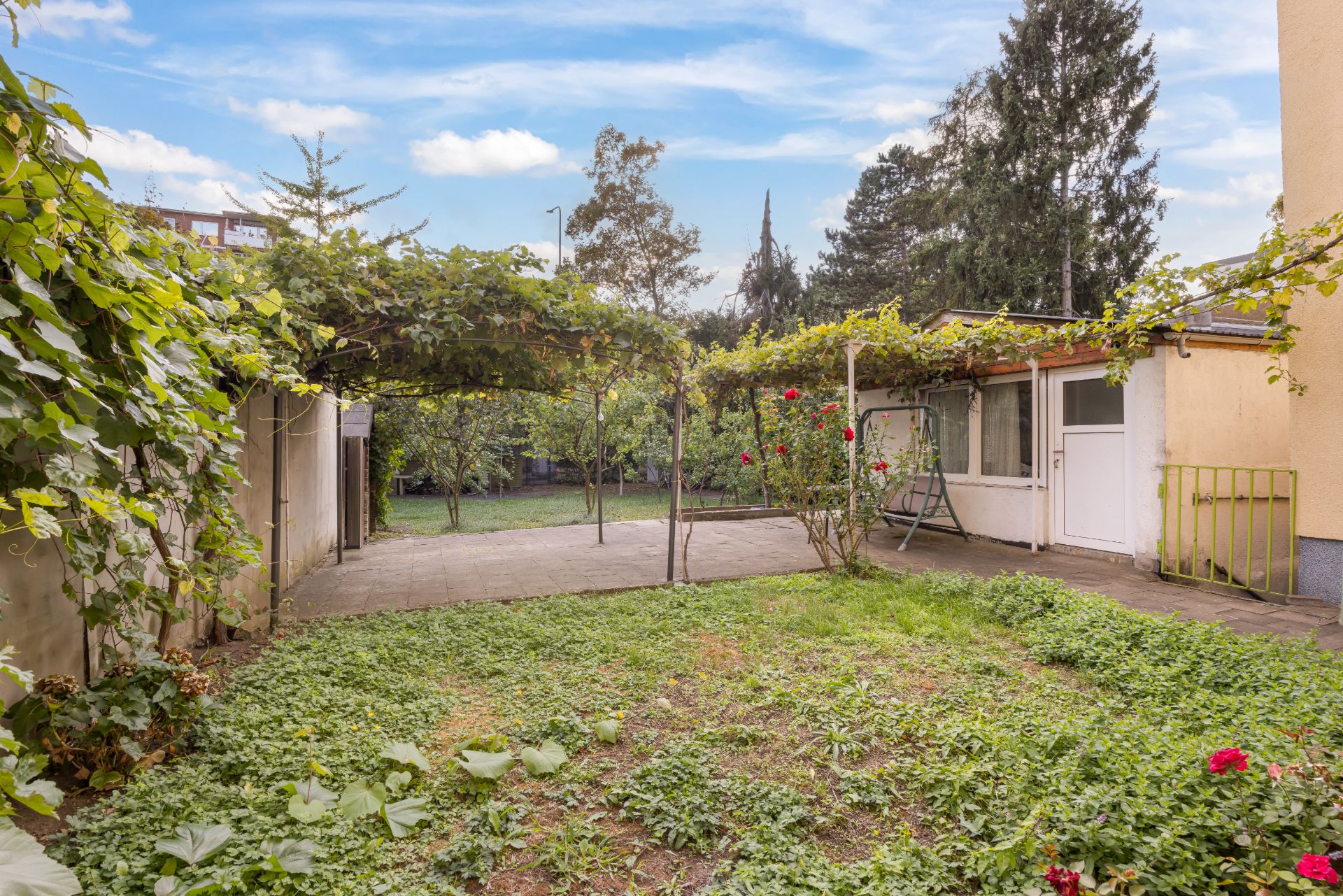 Garten mit überdachter Terrasse, Grünfläche, Kletterpflanzen und Nebengebäude.