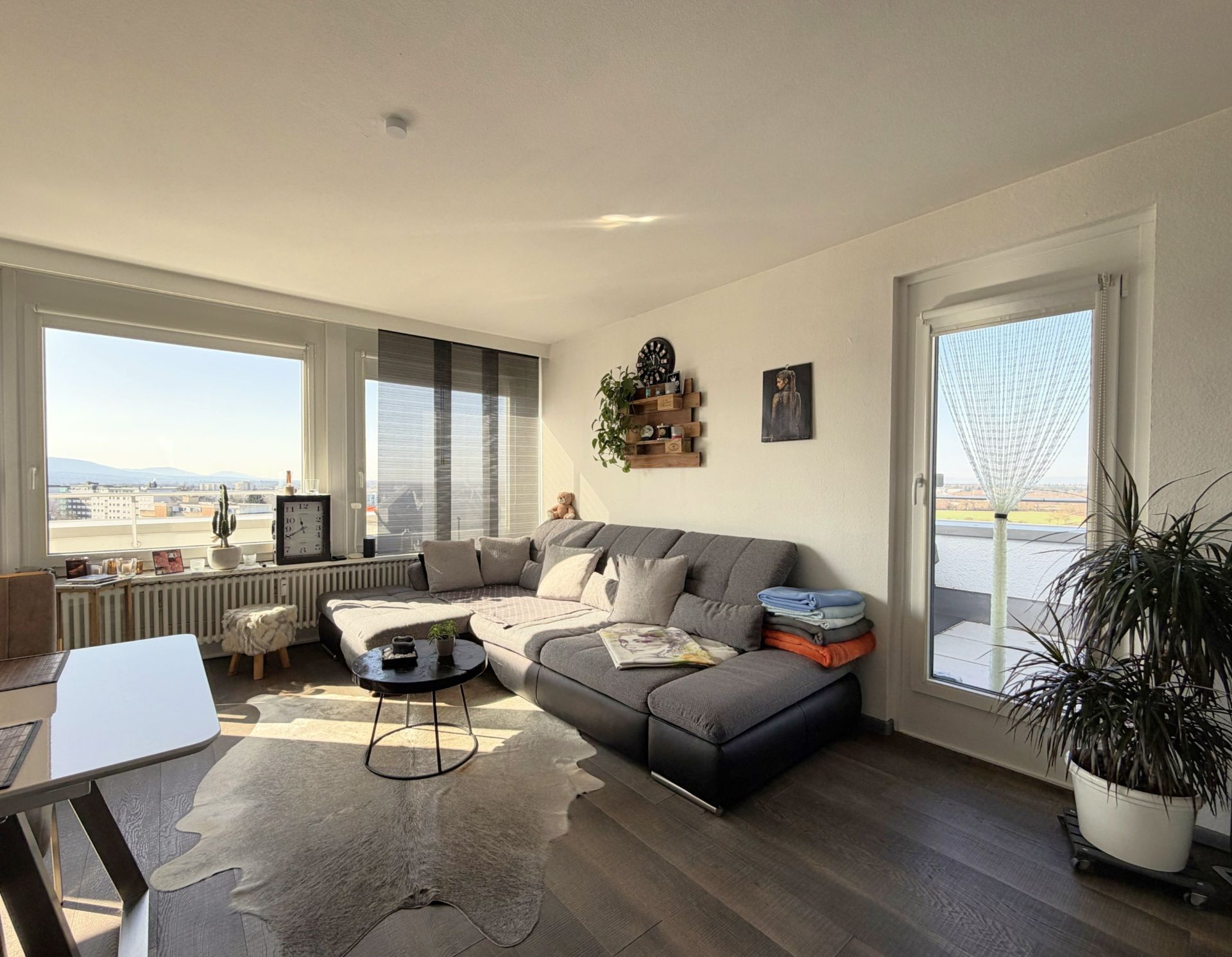 Helles Wohnzimmer mit grauem Ecksofa, großen Fenstern und Zugang zu einem Balkon.