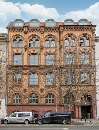 Mehrstöckiges Backsteinhaus mit Rundbogenfenstern, davor geparkte Autos und kahle Bäume.