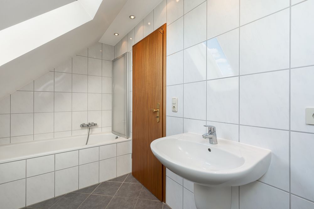Modernes Badezimmer mit weiß gefliester Badewanne, Waschbecken und Holz­tür unter Dachschräge.