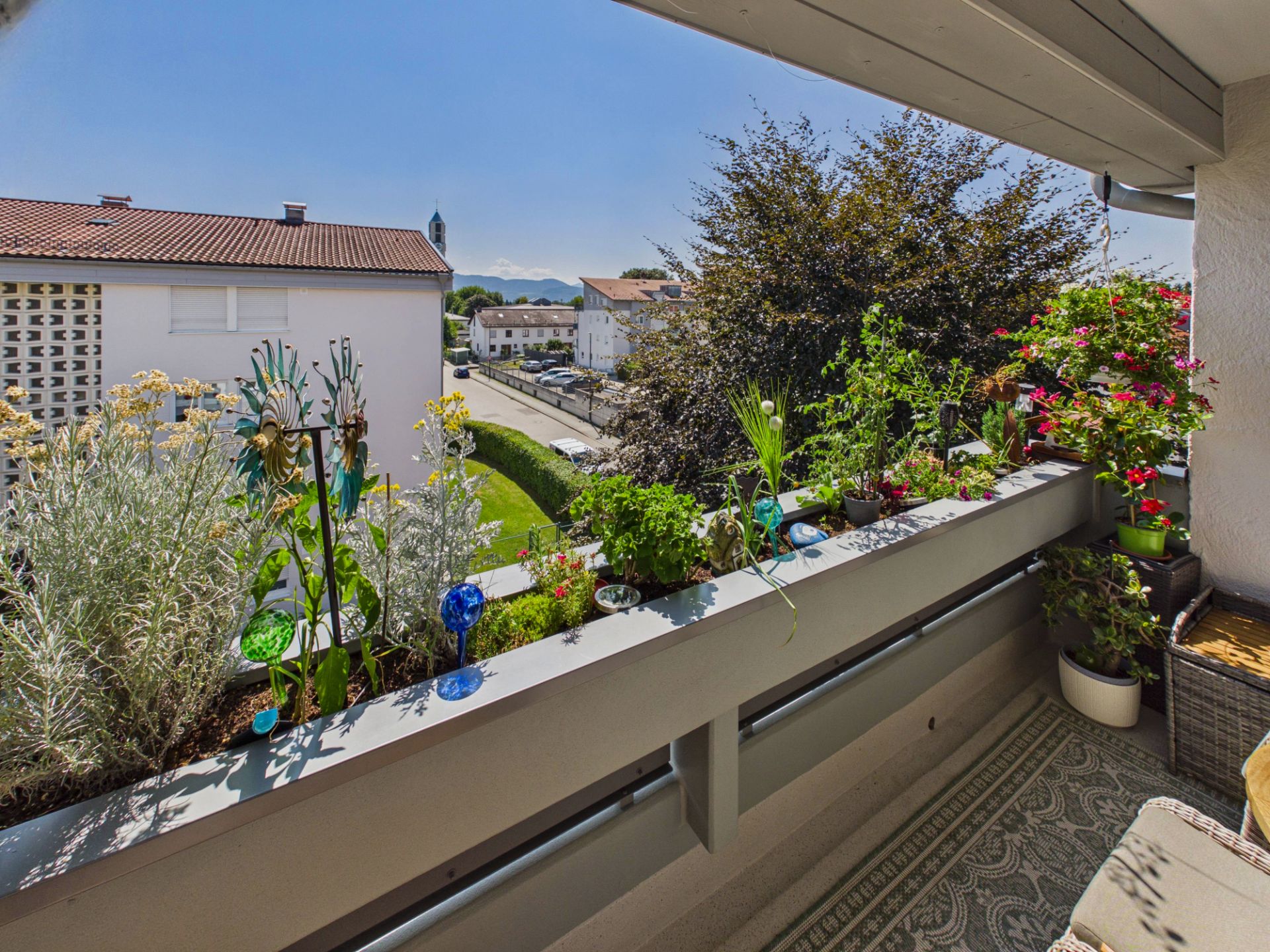 Balkon mit vielen Pflanzen und Blick auf Häuser und Bäume bei sonnigem Wetter.