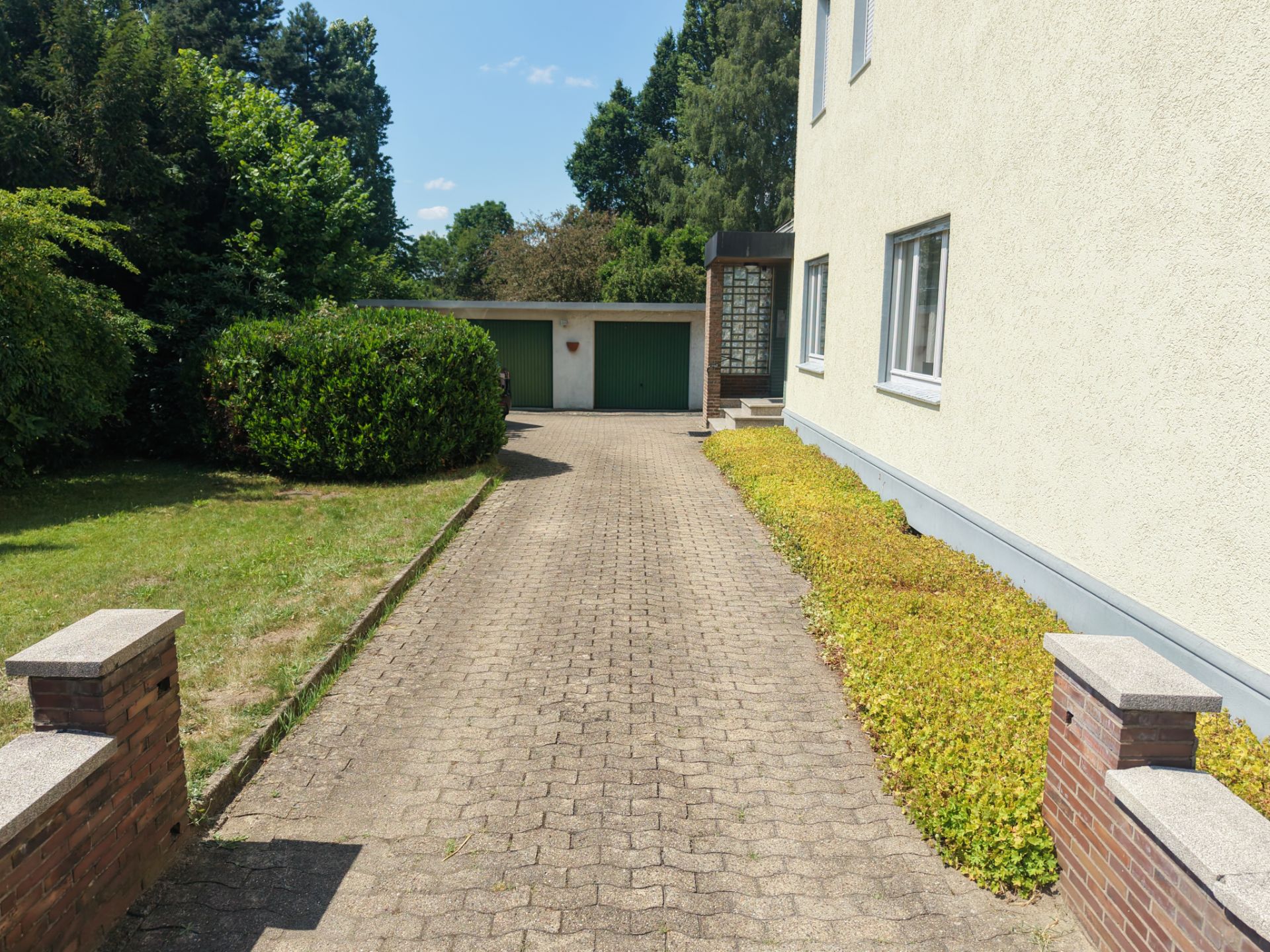 Gepflasterte Einfahrt zu einer Doppelgarage, mit grüner Hecke und Rasen an der Seite.