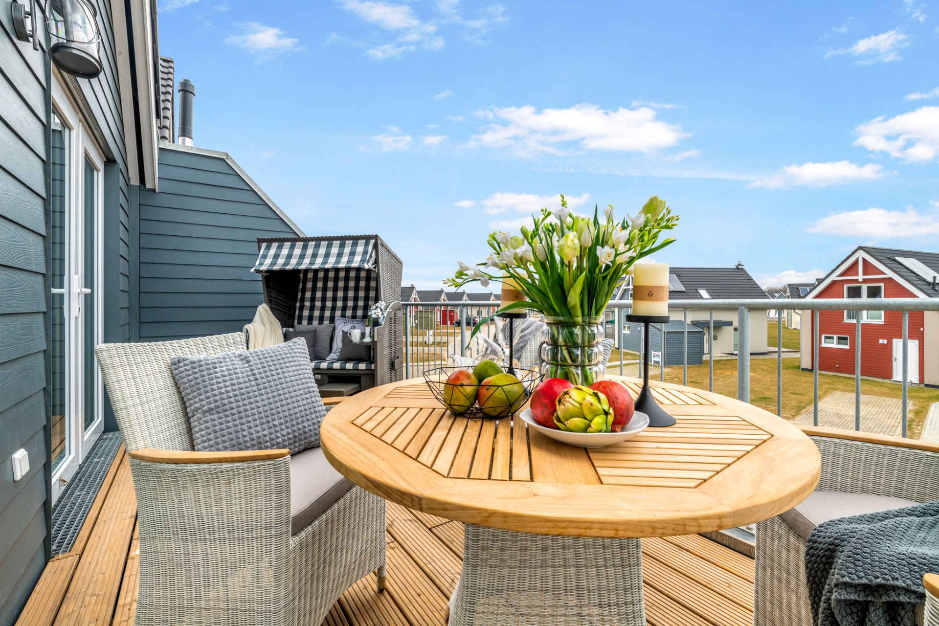 Holzbalkon mit runden Tisch, Obstschale, Blumen und gemütlichen Stühlen, Ausblick auf Nachbarhäuser.