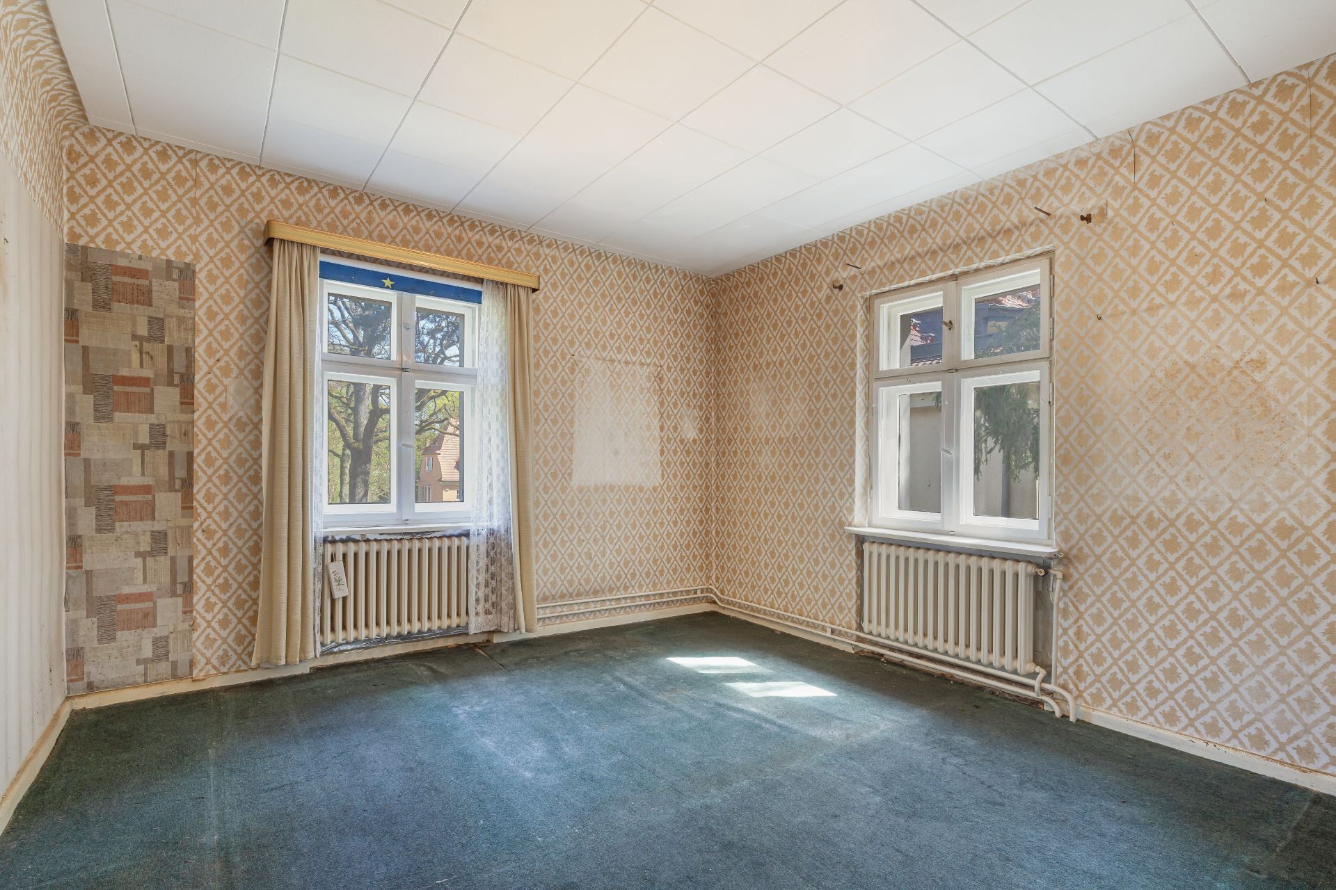 Leerer Raum mit zwei Fenstern, alten Heizkörpern und gemusterter Tapete.