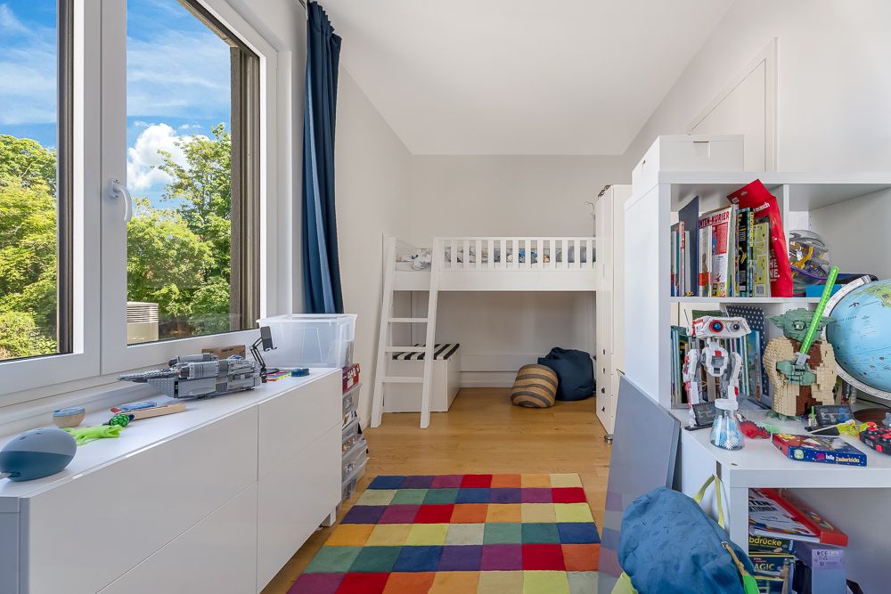 Kinderzimmer mit Hochbett, buntem Teppich, Spielzeug und Büchern, großes Fenster mit Blick ins Grüne.
