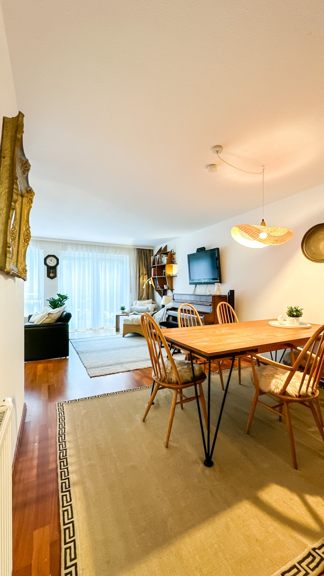 Helles Wohnzimmer mit Esstisch, vier Stühlen, Sofa, Fernseher und großen Fenstern mit Gardinen.