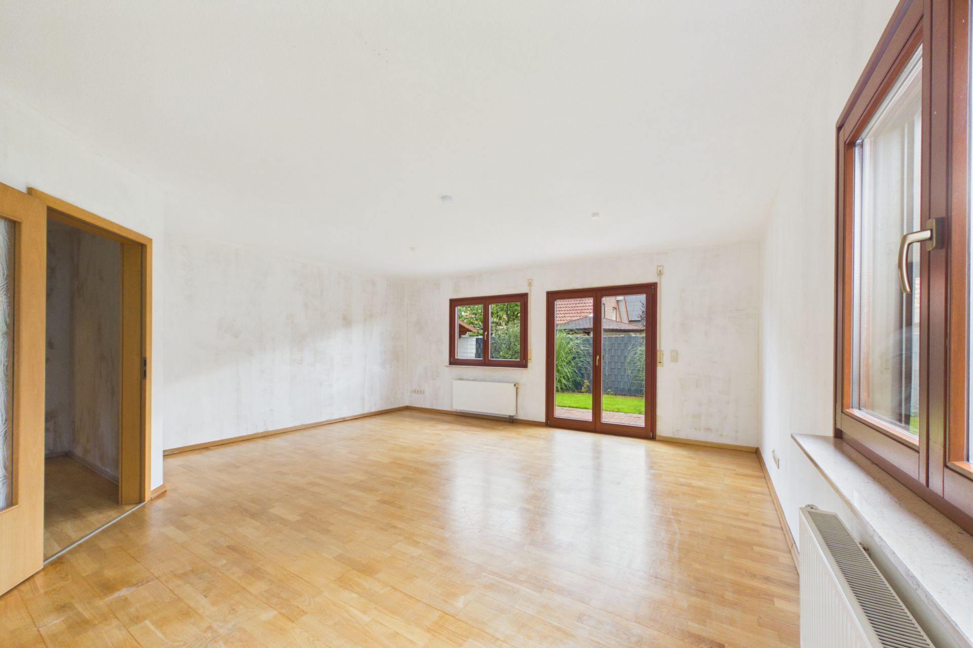 Helles, leeres Wohnzimmer mit Parkettboden, großen Fenstern und Zugang zum Garten.