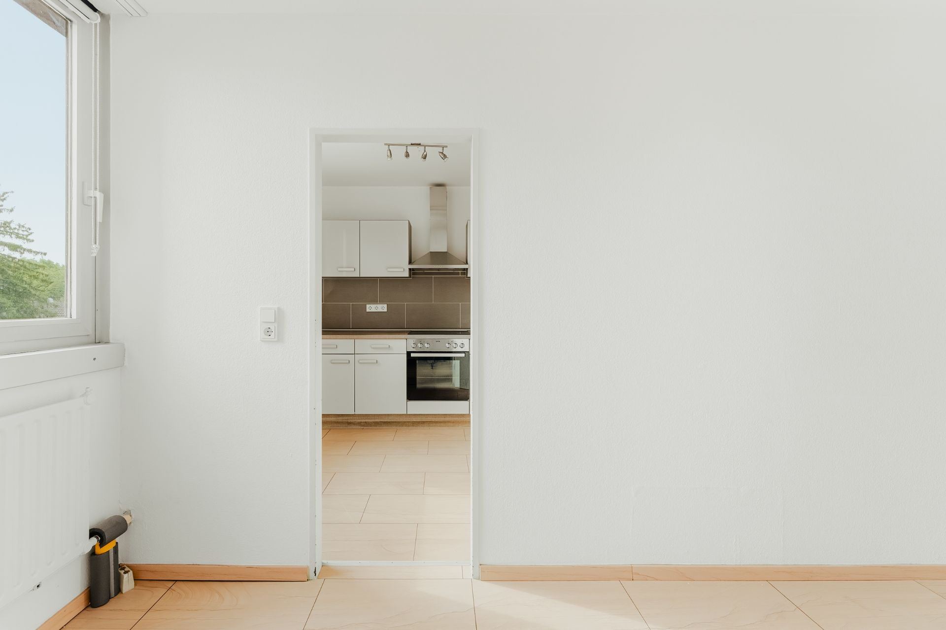 Heller Raum mit Blick durch eine Tür in eine moderne Einbauküche mit weißen Schränken und Backofen.