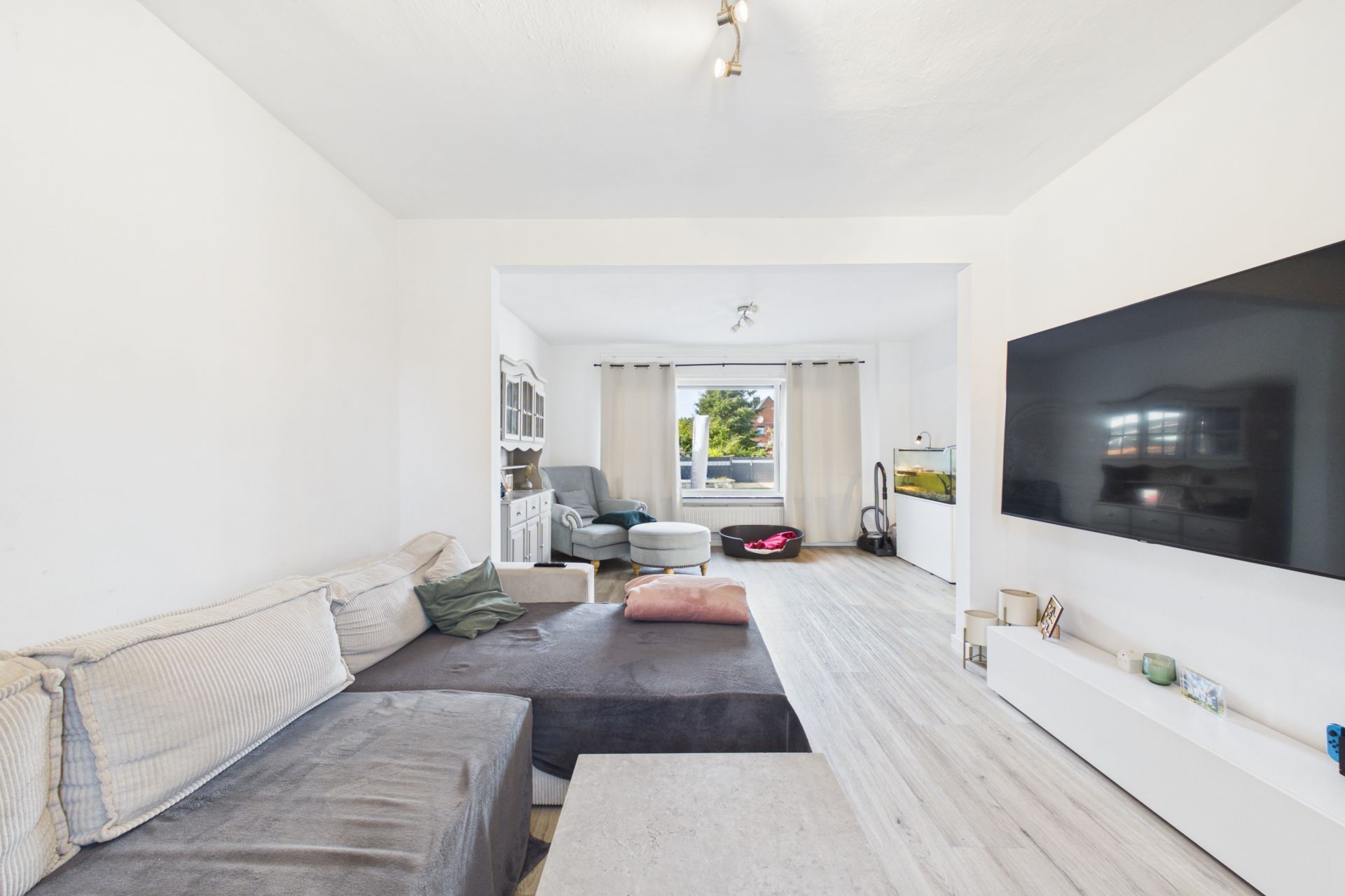 Helles Wohnzimmer mit Ecksofa, großem Fernseher und Fenster mit Blick nach draußen.
