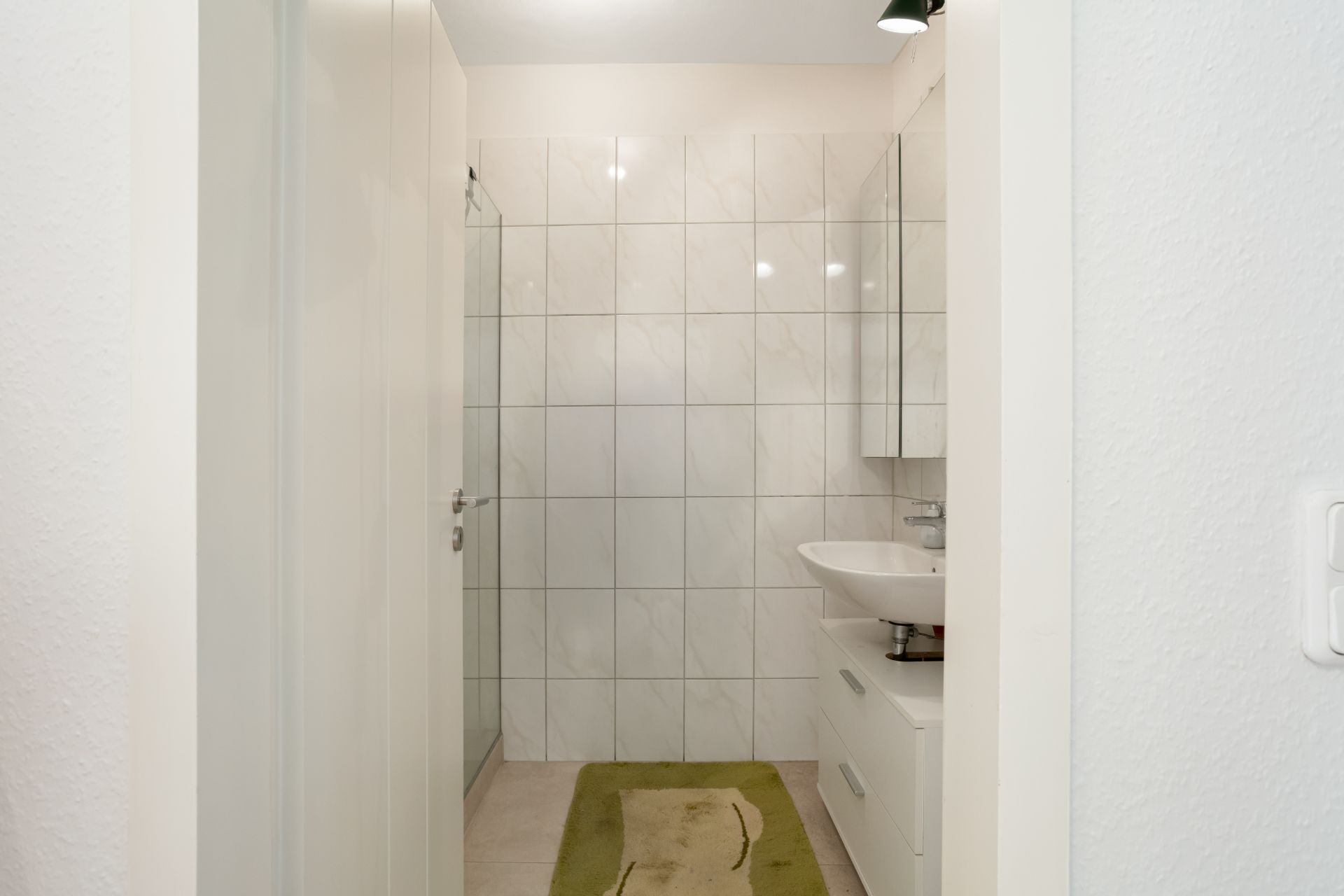 Modernes Badezimmer mit weiß gefliesten Wänden, Waschbecken, Spiegel und grüner Badematte.