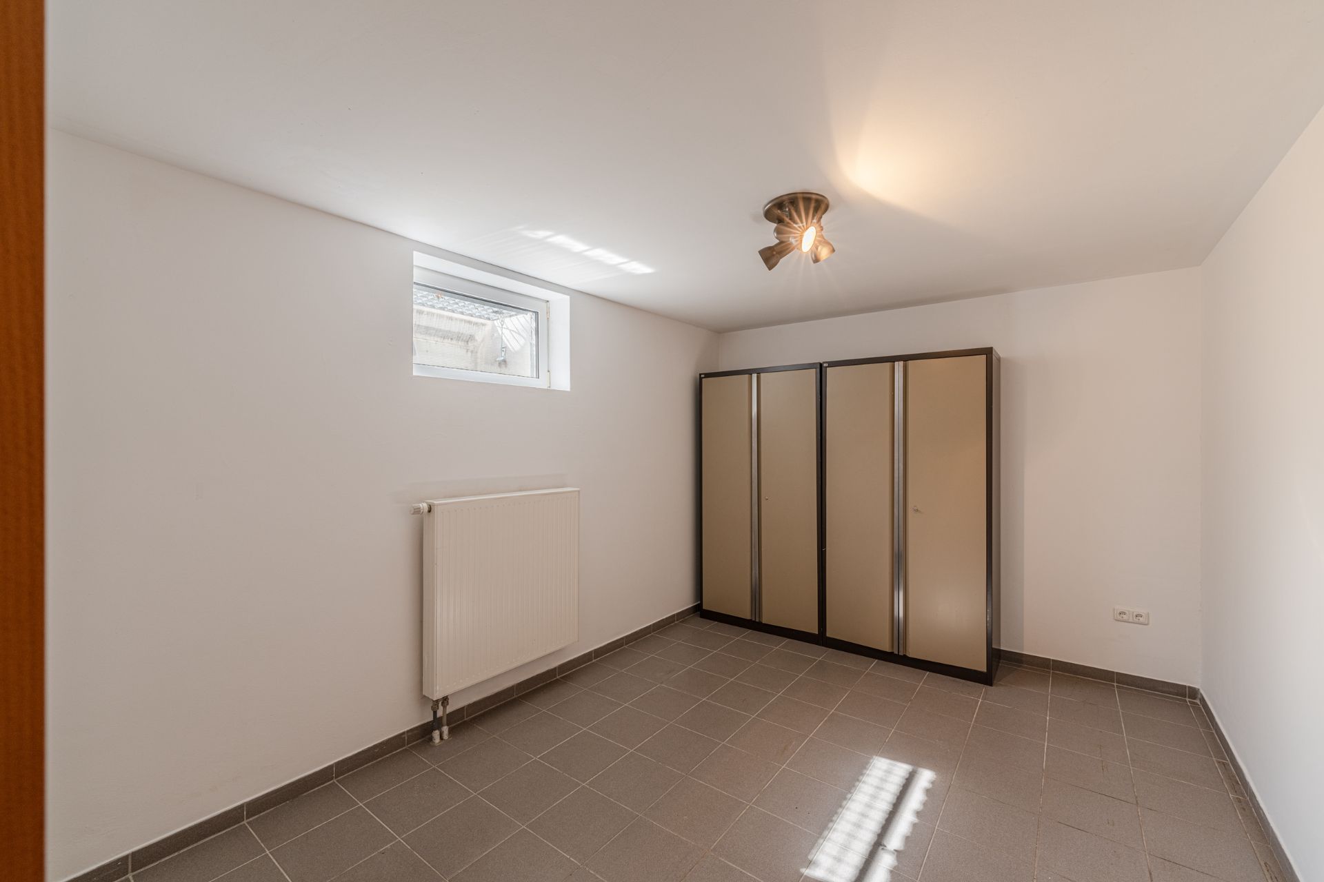 Leerer Raum mit grauen Fliesen, kleinem Fenster, Heizkörper und drei Schränken.