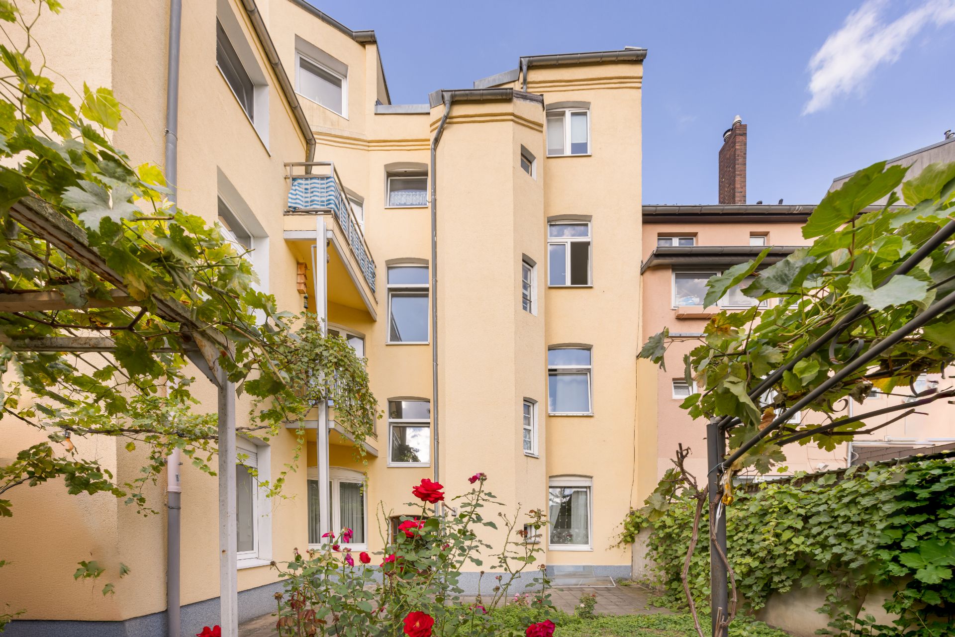 Mehrstöckiges Wohngebäude mit gelber Fassade, Balkon und Garten mit Rosen im Vordergrund.