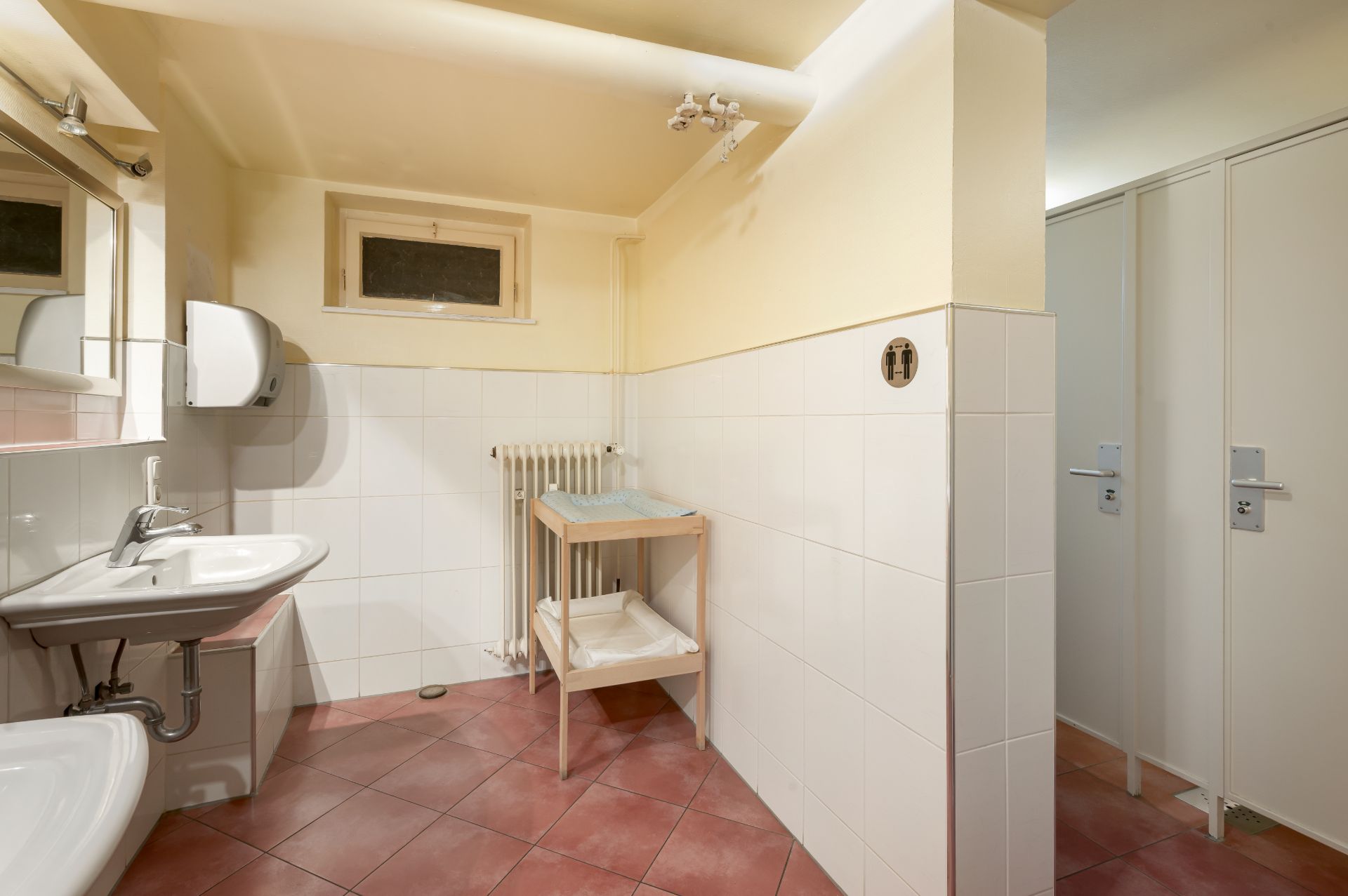 Helles Badezimmer mit Waschbecken, Spiegel, Heizkörper und Türen zu mehreren Kabinen.