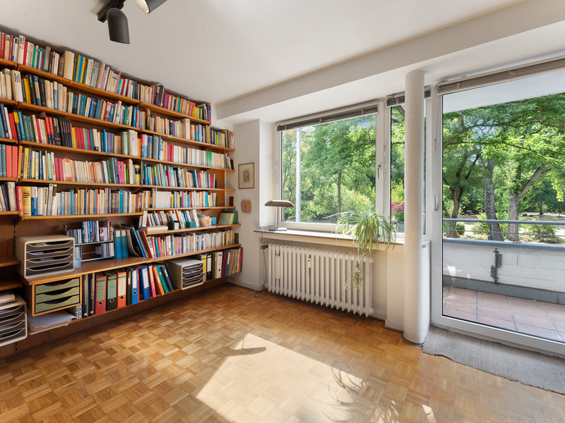 Helles Zimmer mit Bücherregal, großem Fenster, Parkettboden und Blick auf grüne Bäume.