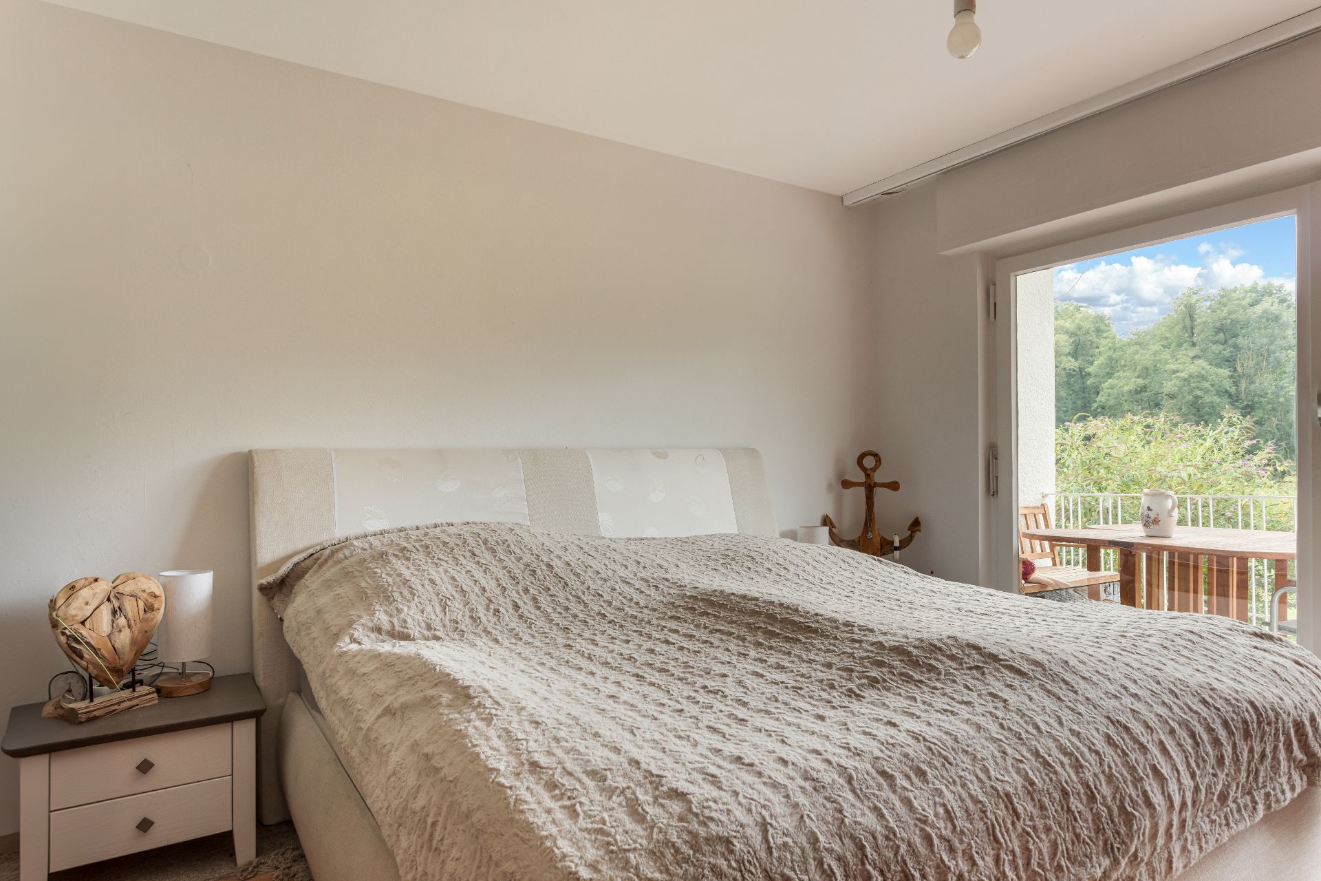 Schlafzimmer mit Doppelbett, Nachttisch und Zugang zu Balkon mit Holztisch und Stühlen.