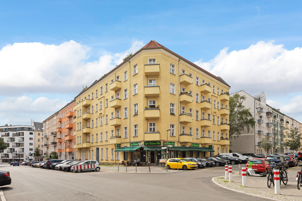 Ecke eines gelben Wohnhauses mit Balkonen, parkenden Autos und blauem Himmel.