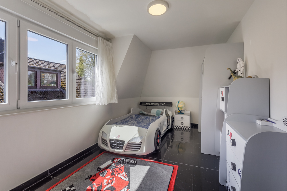 Kinderzimmer mit Auto-Bett, Spielteppich, Fenster und weißen Möbeln.