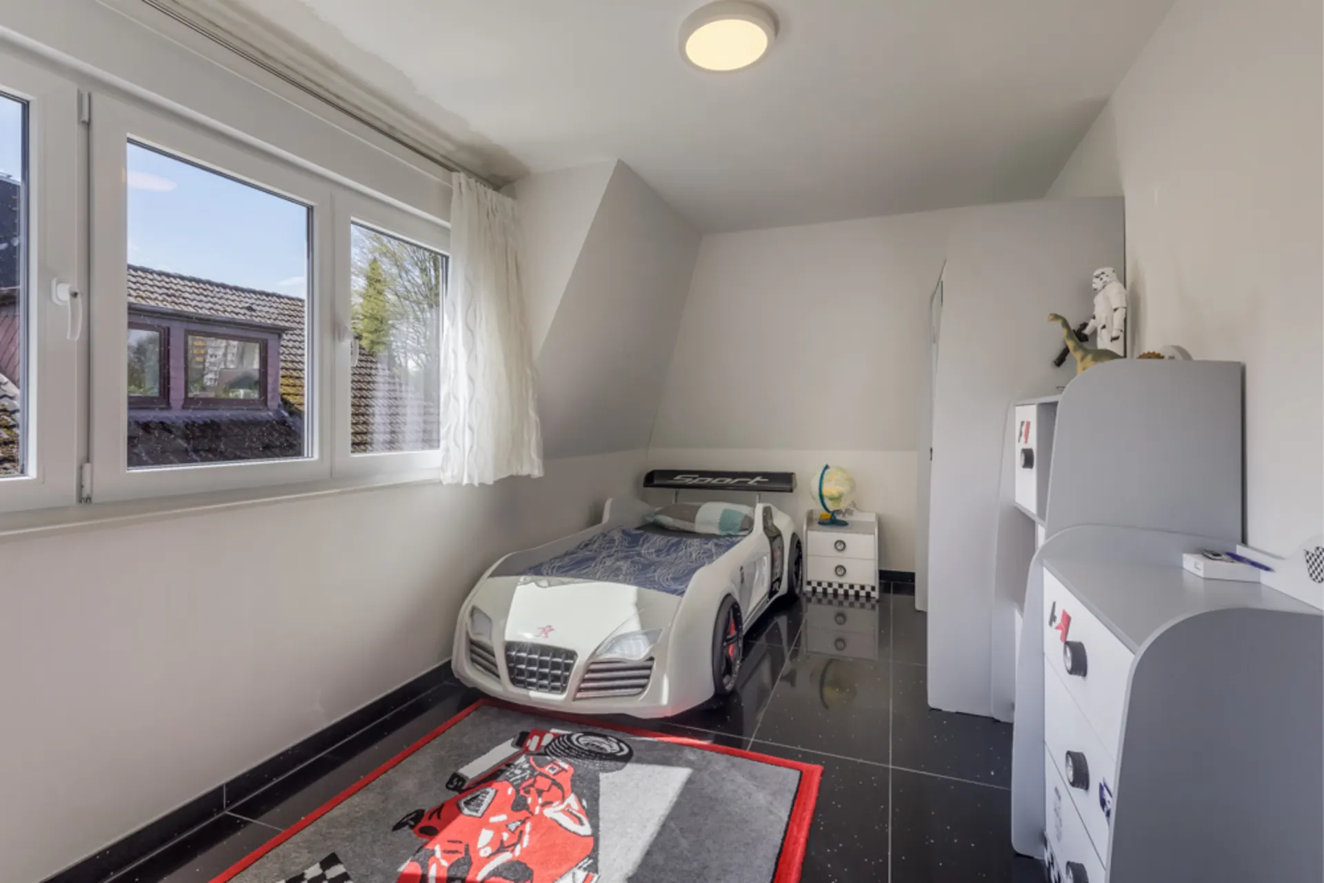 Kinderzimmer mit Auto-Bett, Spielteppich, Fenster und weißen Möbeln.