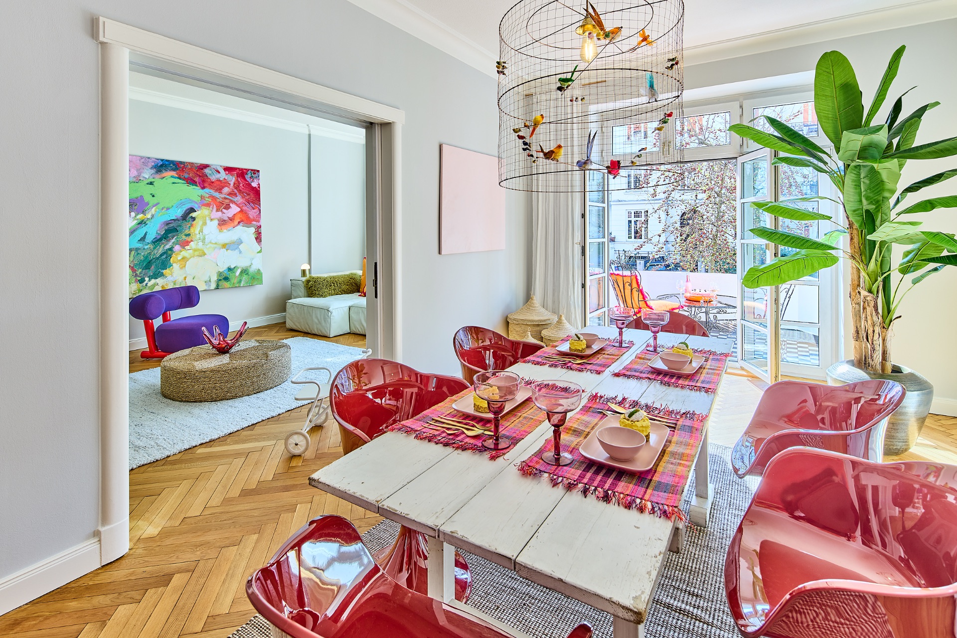 Esszimmer mit weißem Tisch, roten Stühlen, großer Pflanze und Blick in buntes Wohnzimmer.