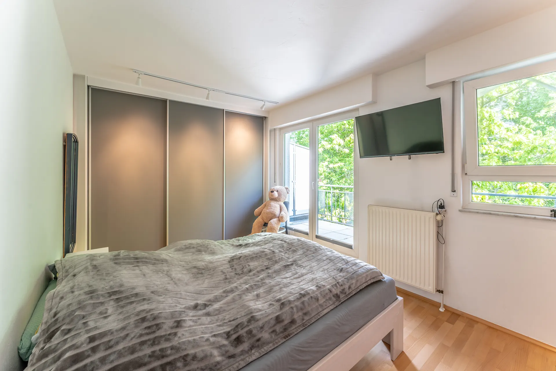 Schlafzimmer mit Doppelbett, grauem Schrank, Fernseher und großem Fenster mit Blick ins Grüne.