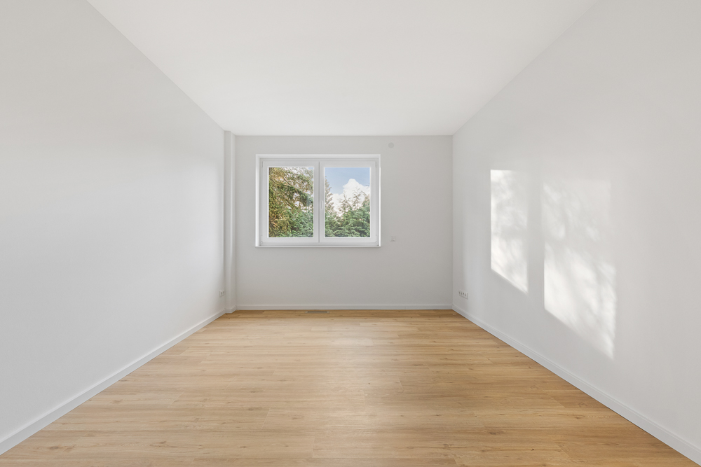 Heller, leerer Raum mit Holzfußboden und einem Fenster, durch das Bäume zu sehen sind.