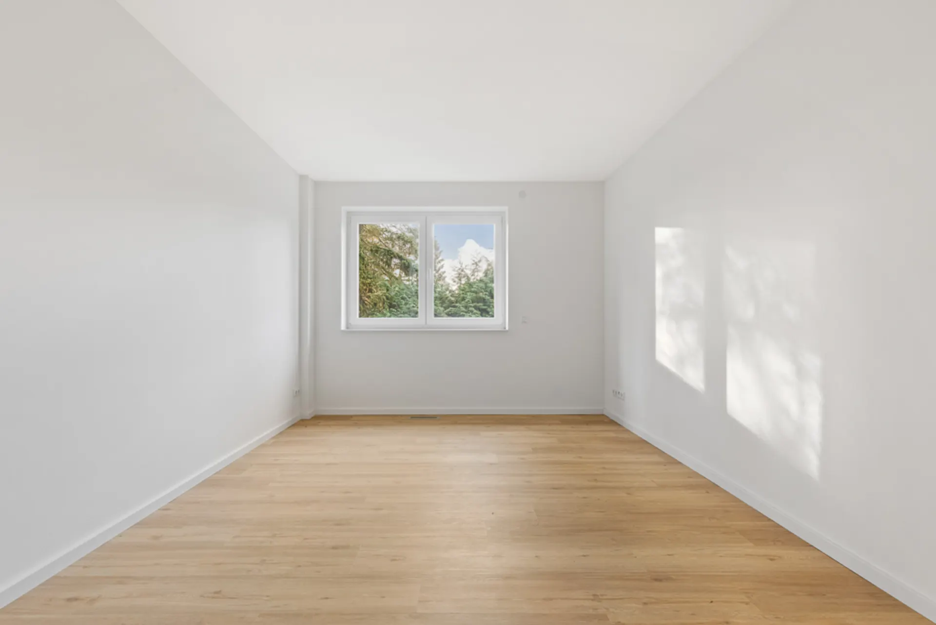 Heller, leerer Raum mit Holzfußboden und einem Fenster, durch das Bäume zu sehen sind.
