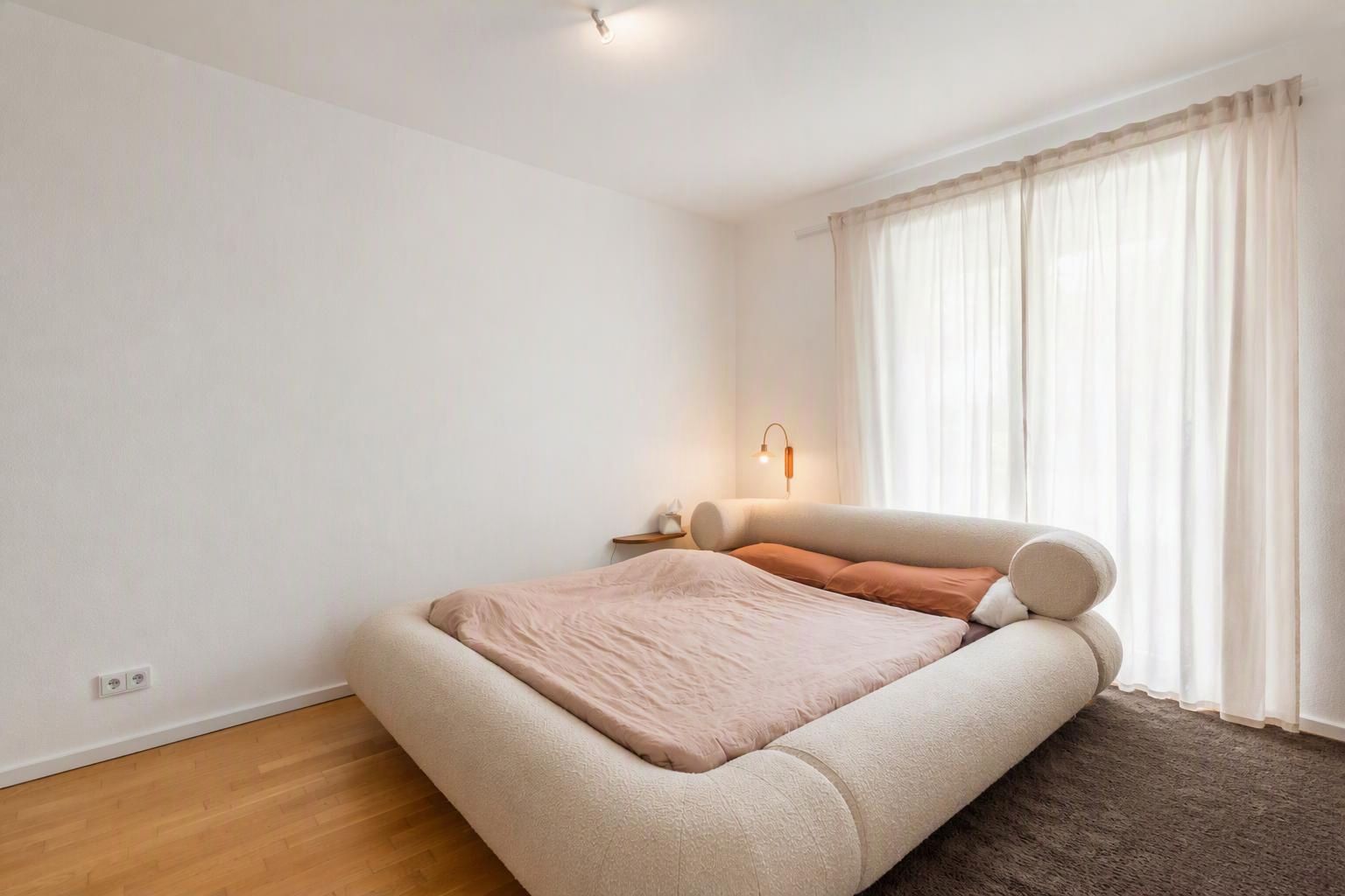 Helles Schlafzimmer mit großem Bett, Nachttisch und bodentiefem Fenster mit Vorhängen.