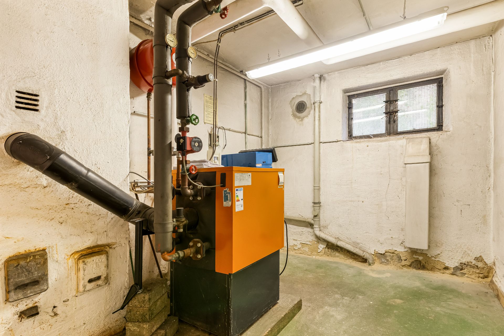 Heizungsraum mit orangefarbener Heizungsanlage, Rohren und kleinem Fenster.