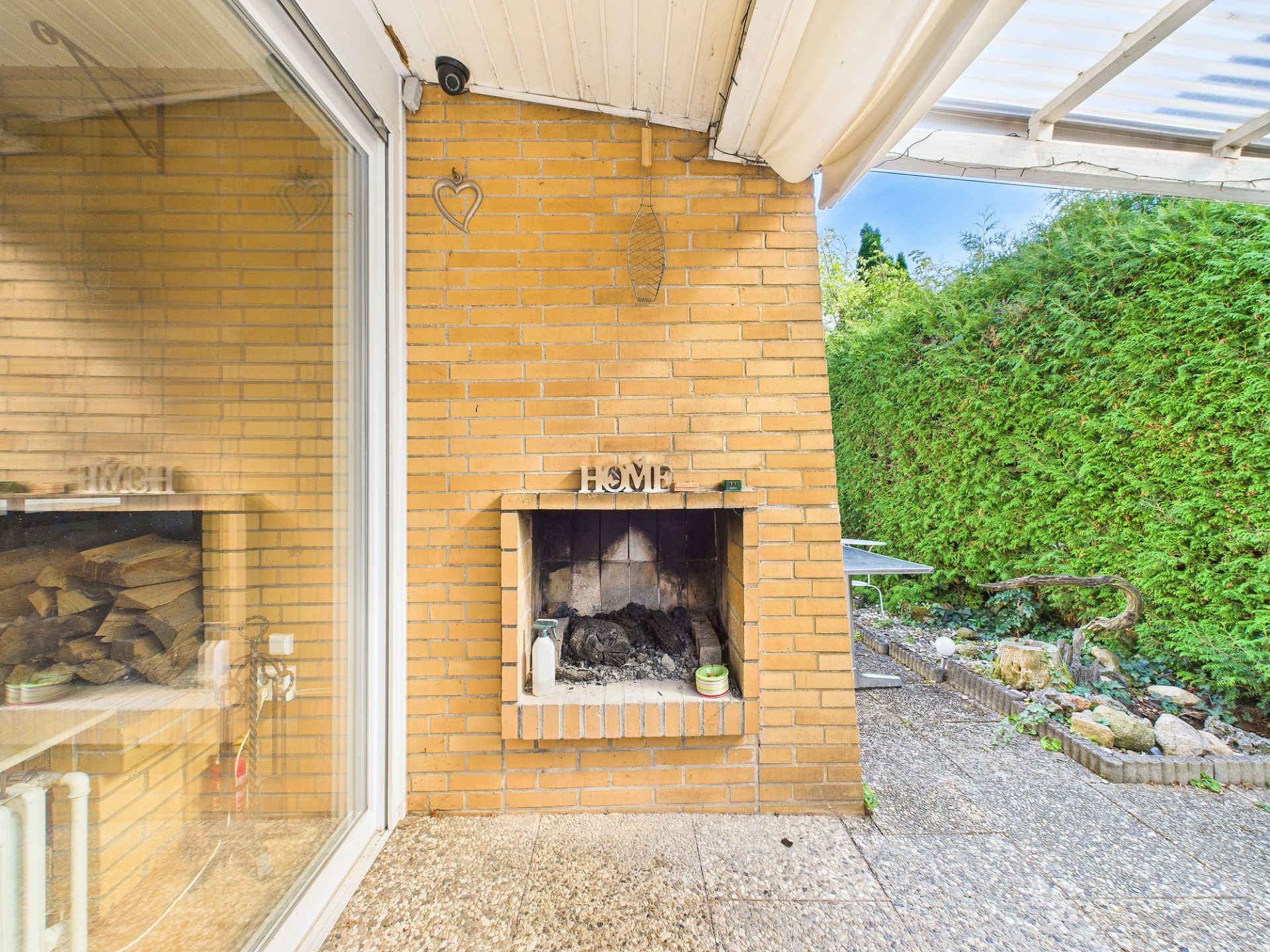 Offener gemauerter Kamin auf einer Terrasse mit Blick auf Garten und Hecke.