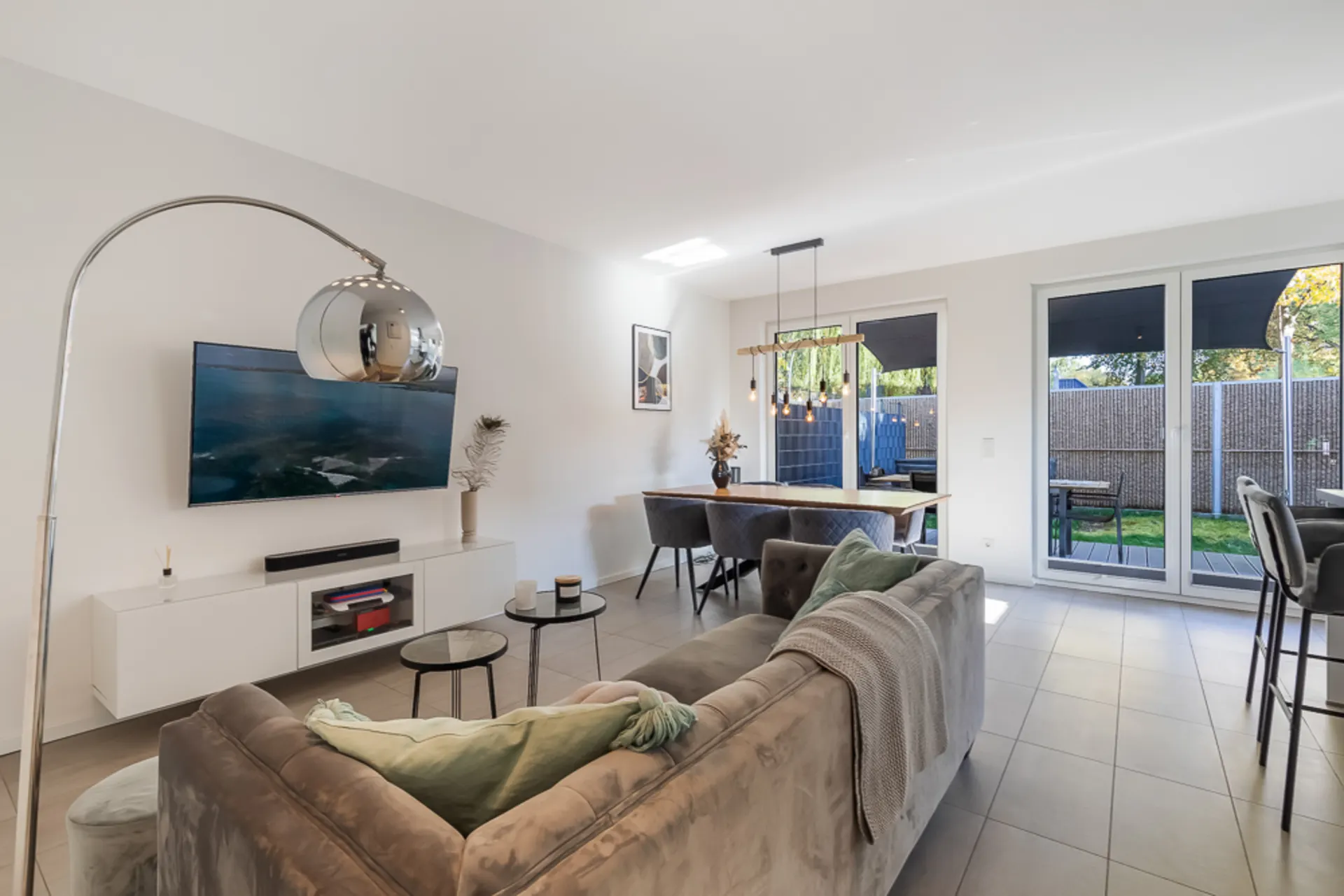 Helles Wohnzimmer mit Sofa, Fernseher, Esstisch und Zugang zur Terrasse mit Garten.